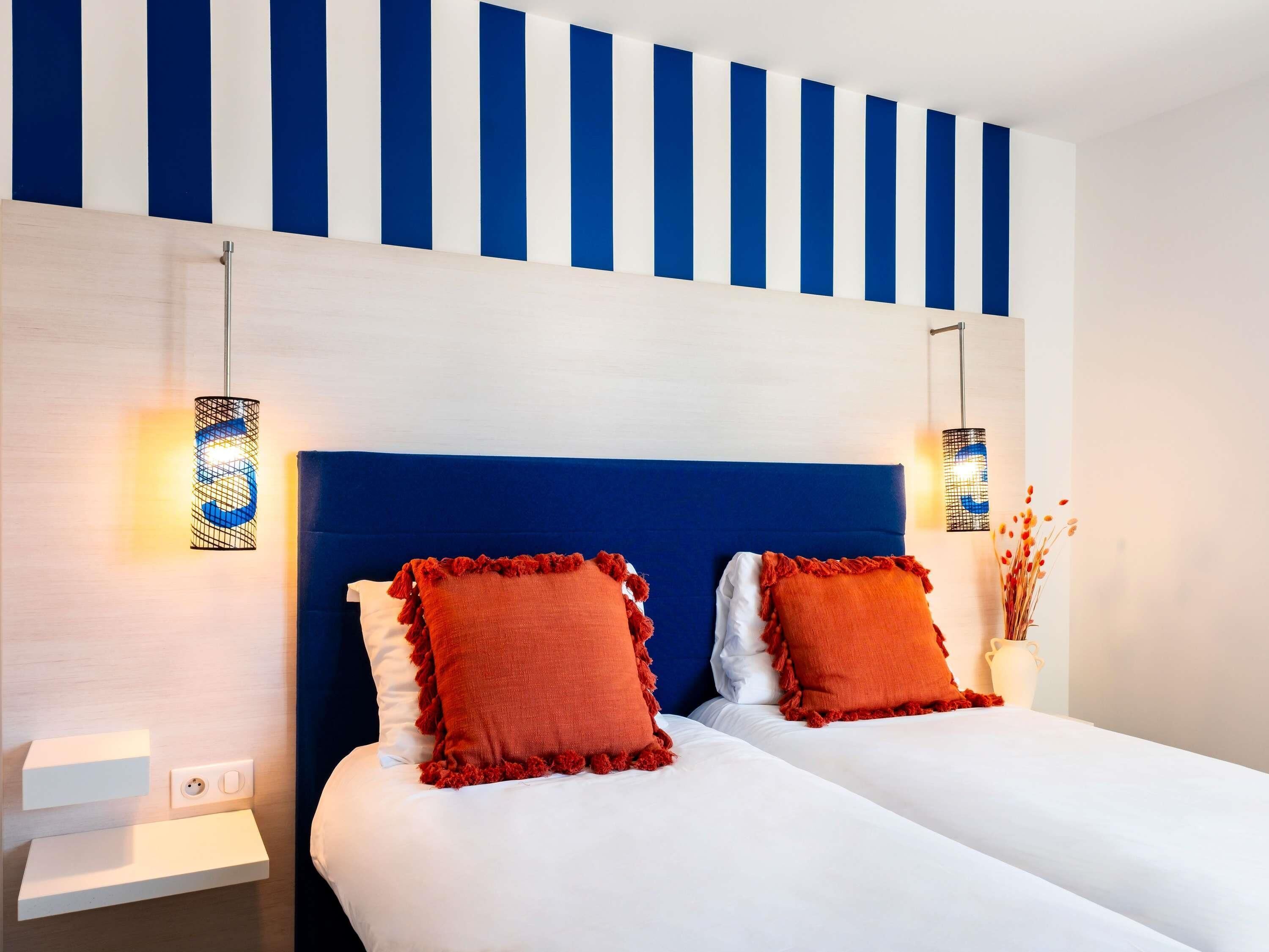 Habitación de hotel con cabecera azul, dos almohadas rojas, ropa de cama blanca y una lámpara de pared.