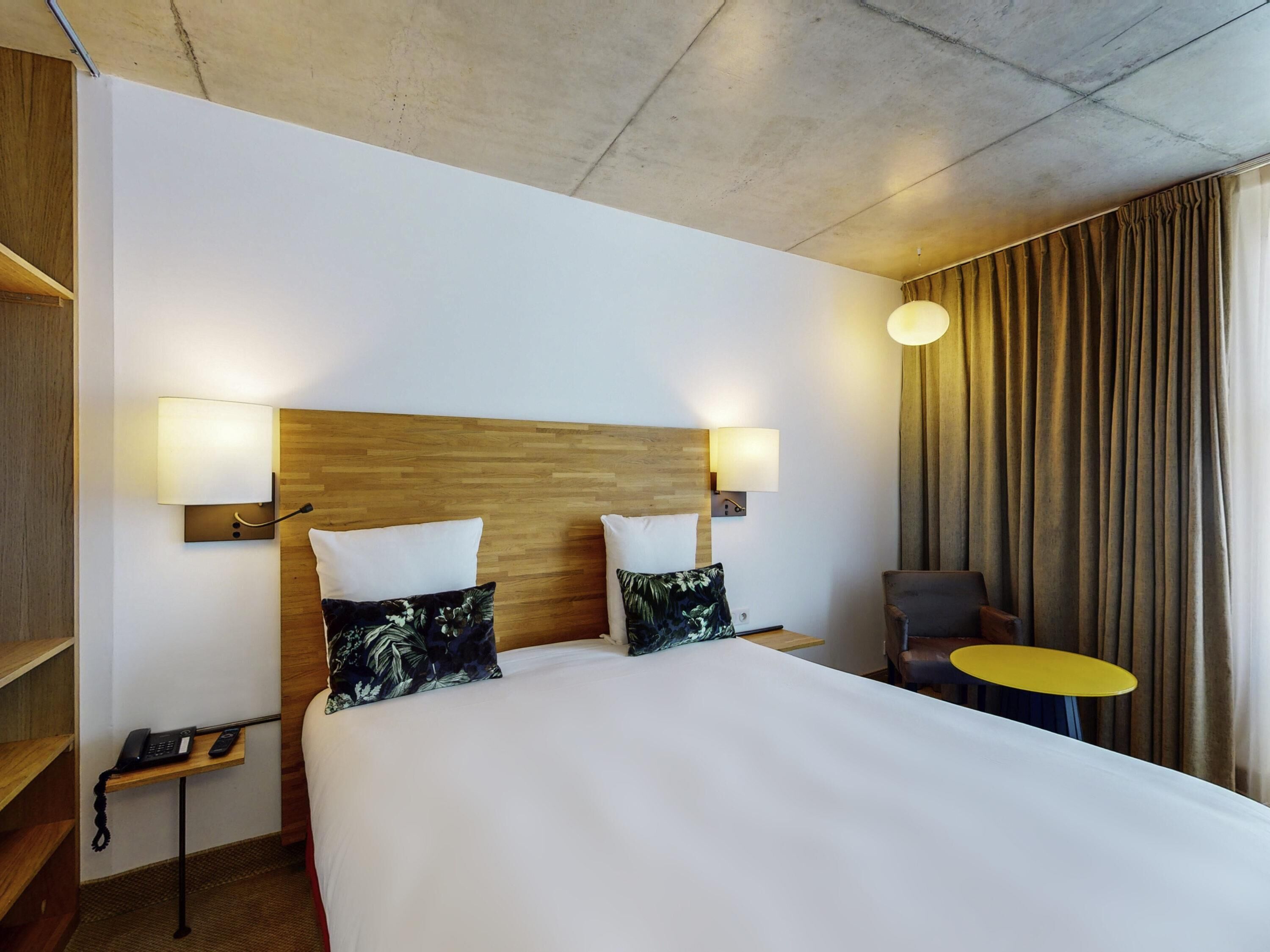 Een hotelkamer met een groot bed, twee bedlampen, een klein tafeltje, een stoel en een gordijn.