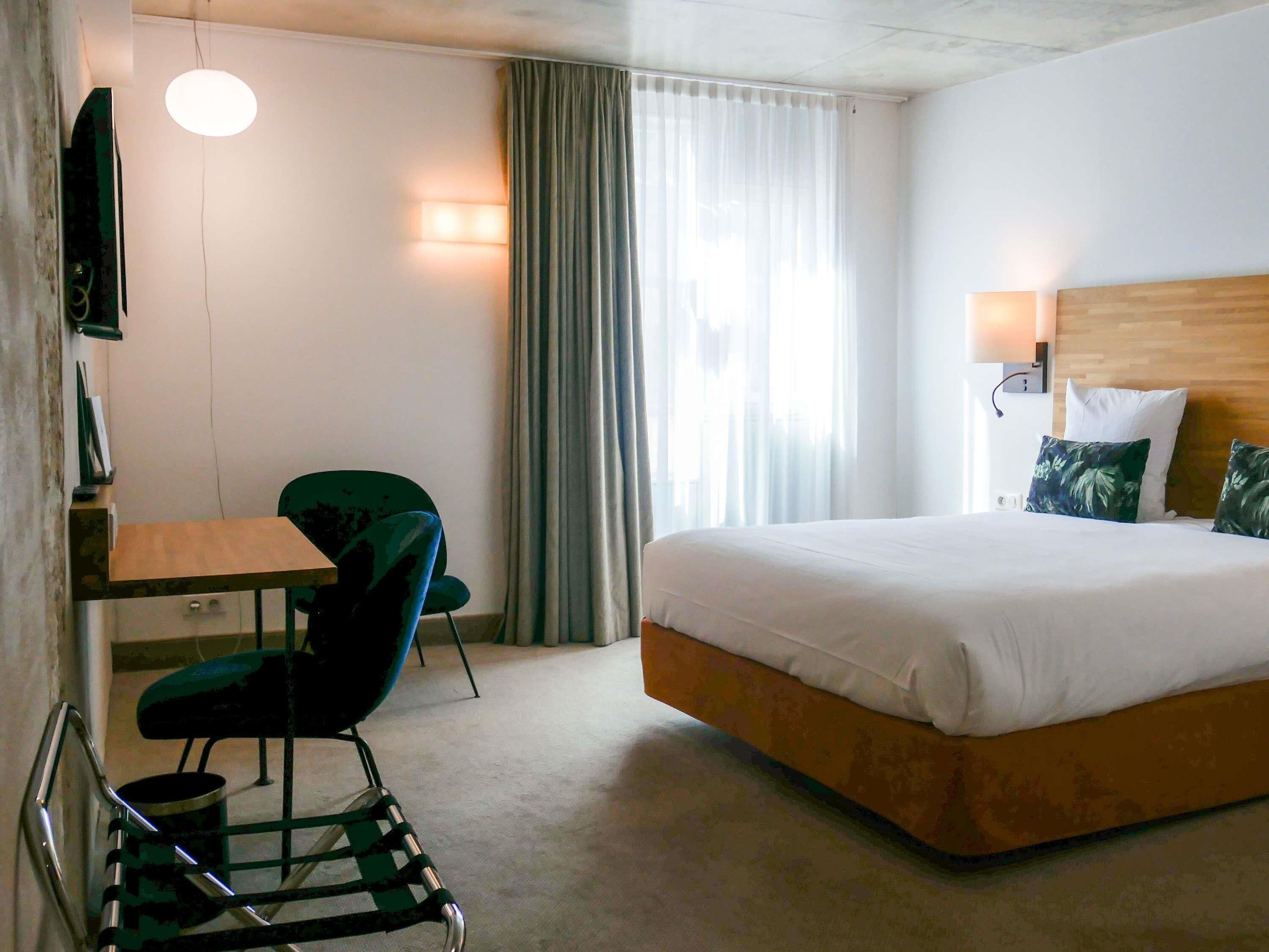 Una habitación de hotel con cama, escritorio, silla, televisor y ventana con cortinas.