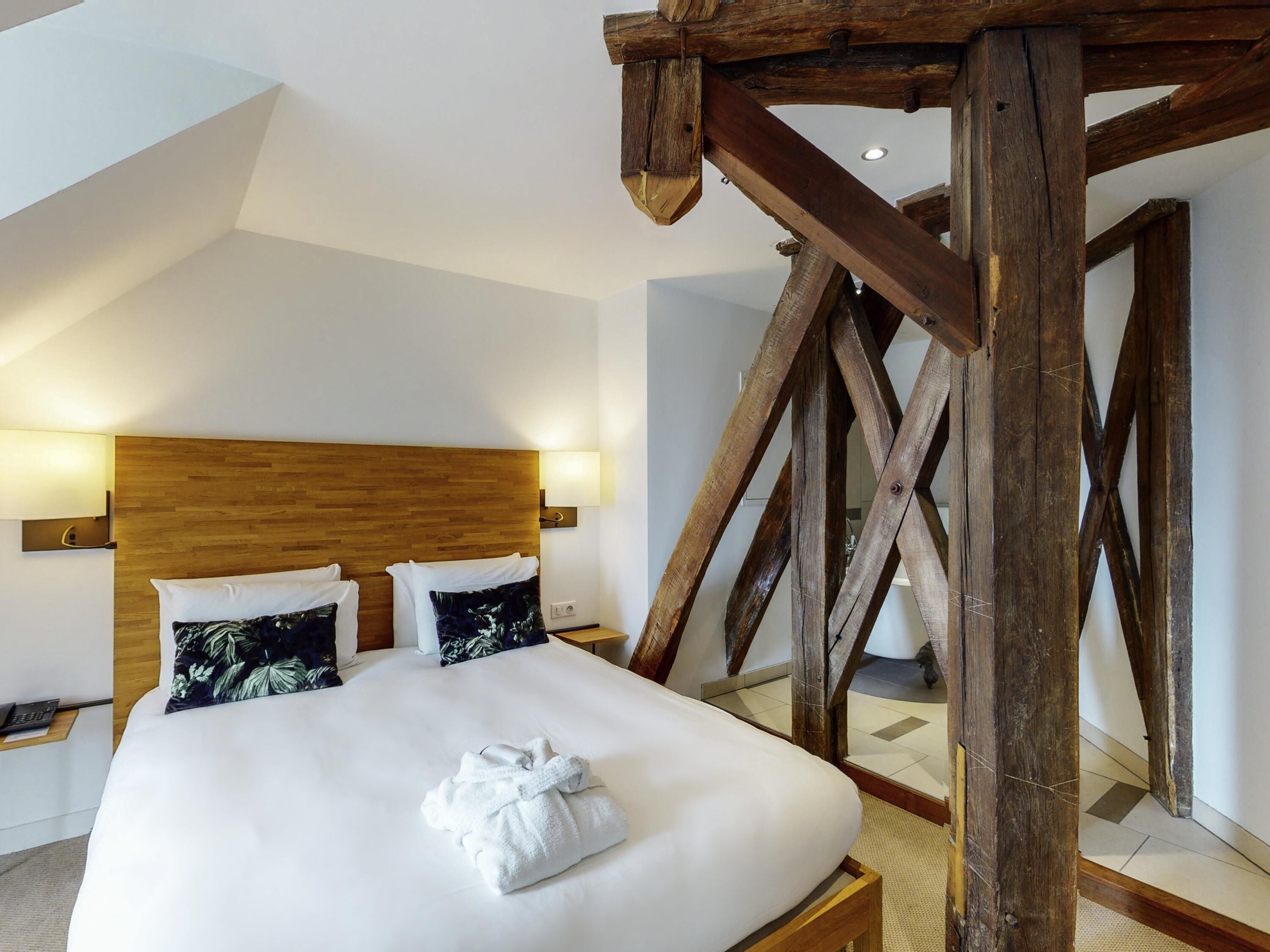 A hotel room with a wooden ceiling structure, a bed with white bedding, and two wall-mounted lamps.