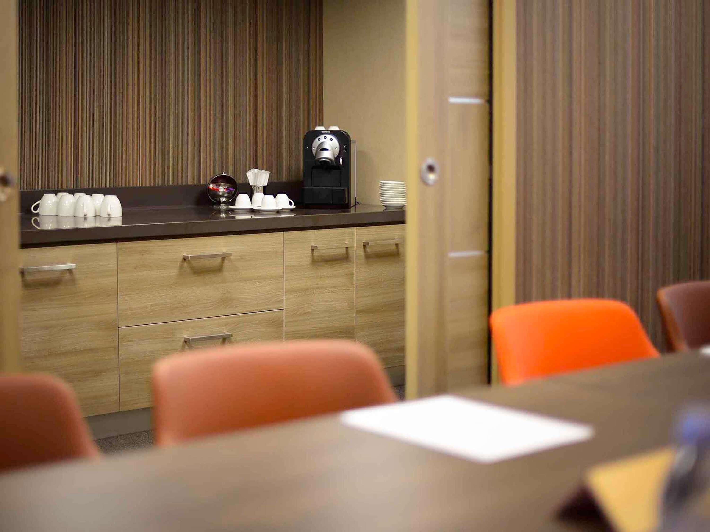 A conference room with a wooden table, orange chairs, a coffee machine, and white cups on the counter.