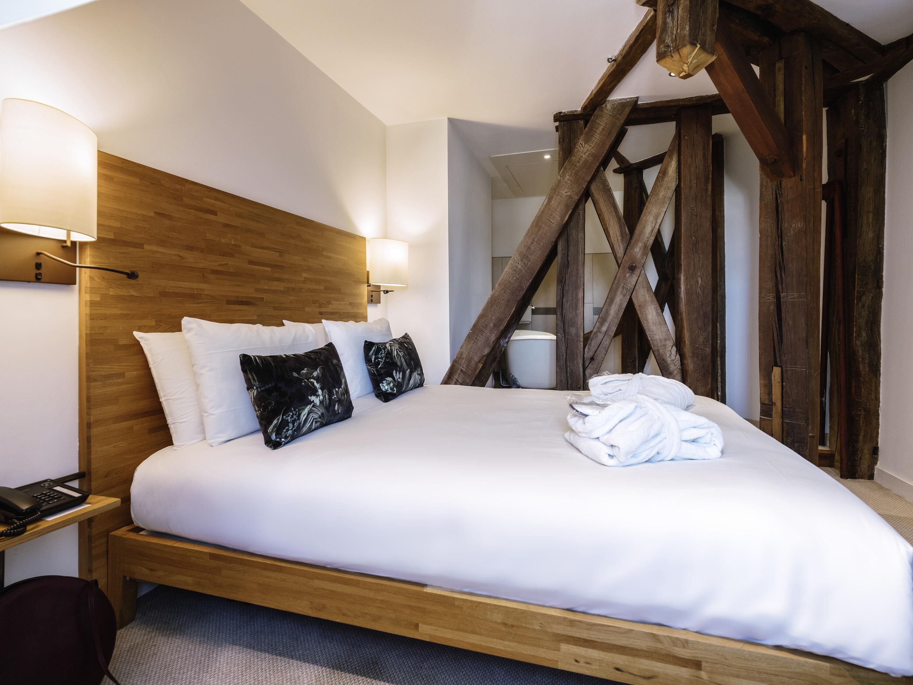 Quarto de hotel com cabeceira de madeira, roupa de cama branca, telefone de apoio e teto de vigas de madeira.