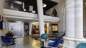 A spacious lobby with a high ceiling, a central column, and a mezzanine level with a railing. There are seating areas with chairs and a small table.