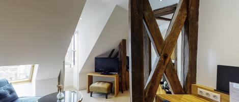 A room with a sloped ceiling, a wooden beam structure, a sofa, a small round table, a television, and a wooden desk with a chair.