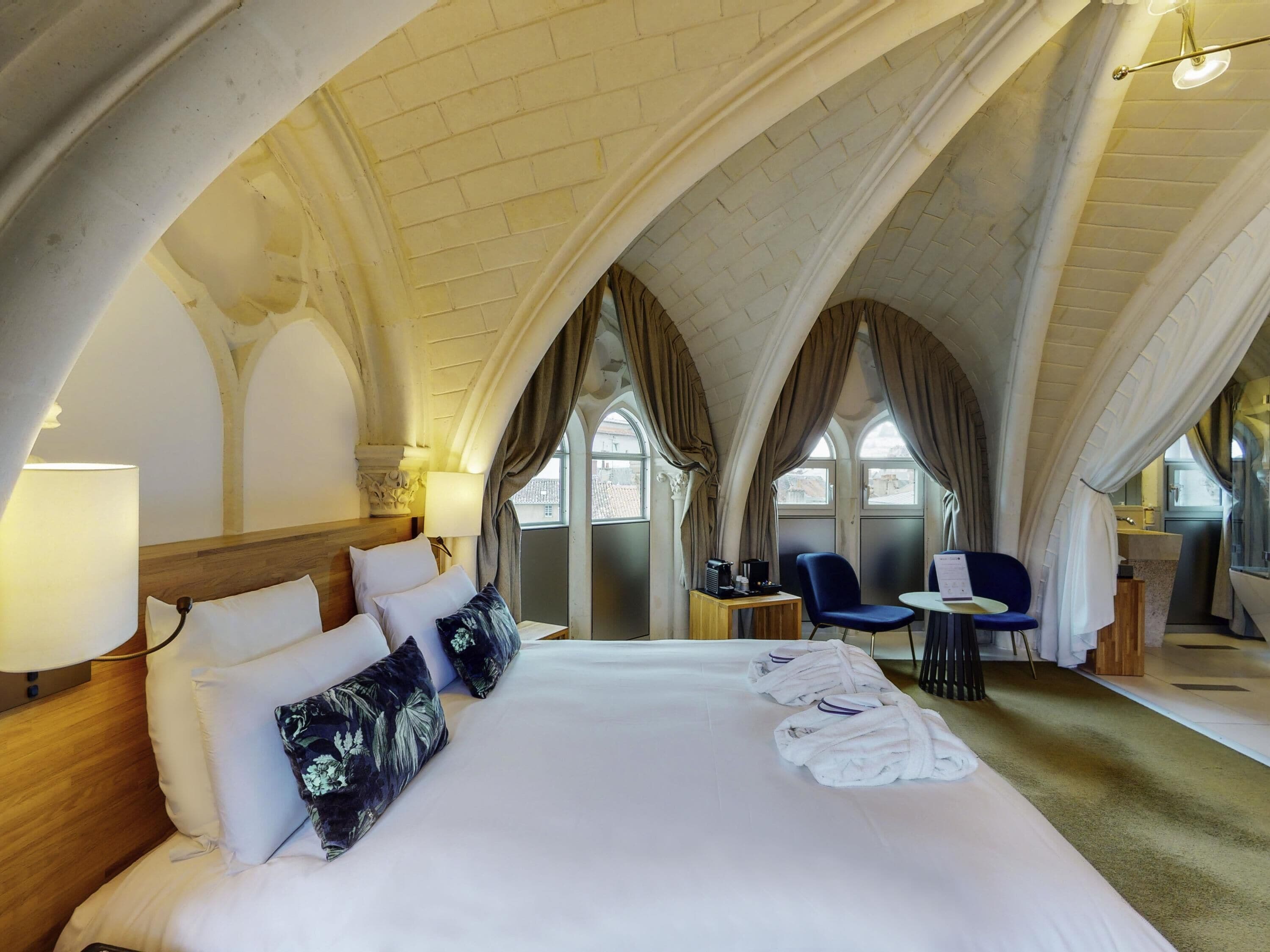 Une chambre d’hôtel avec un grand lit, une petite table, une chaise et une vue sur la ville grâce à de grandes fenêtres en arc de cercle.