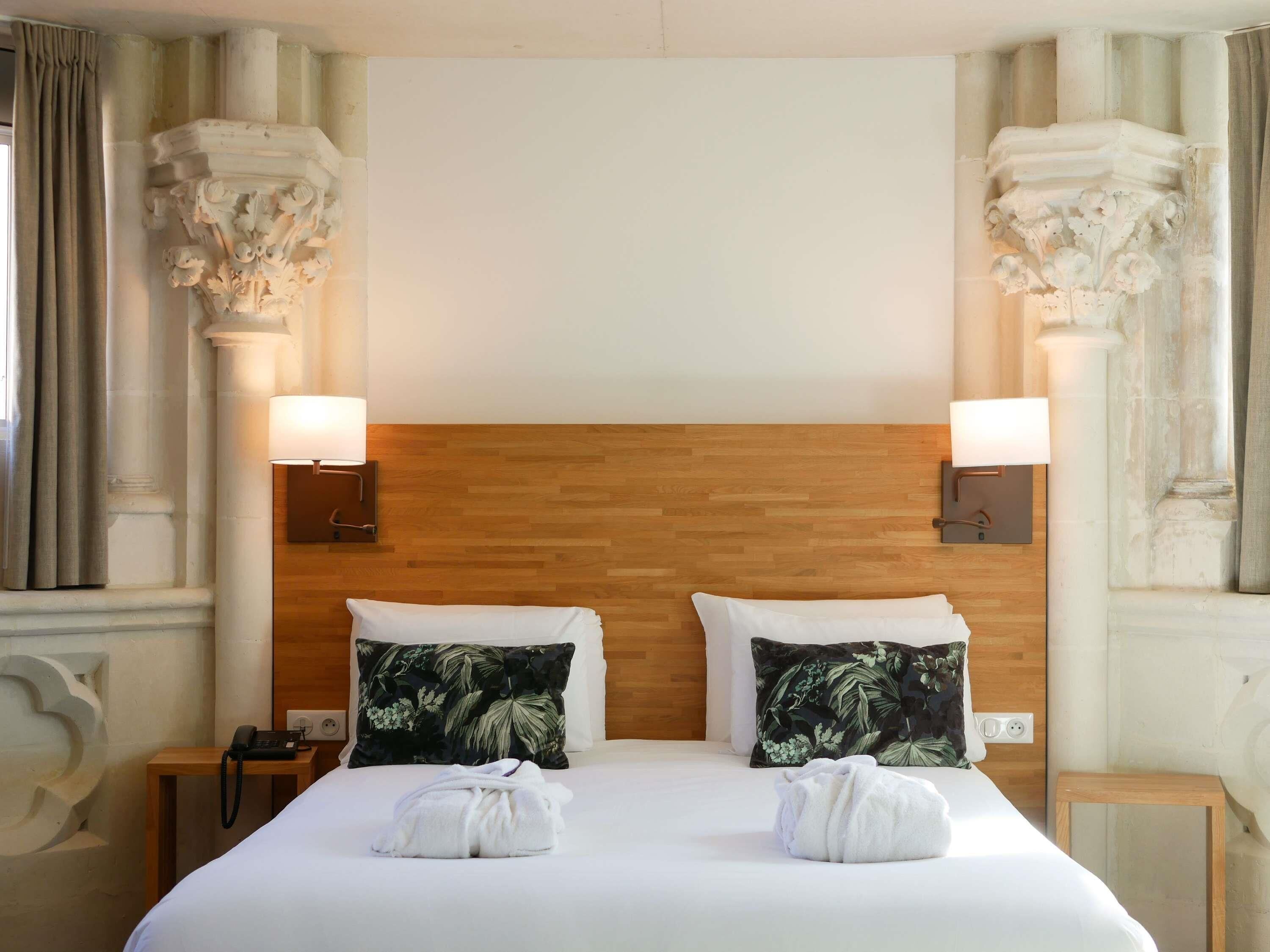 A hotel room with a wooden headboard, two floral pillows, a white bedspread, and a bedside table with a phone and lamp.