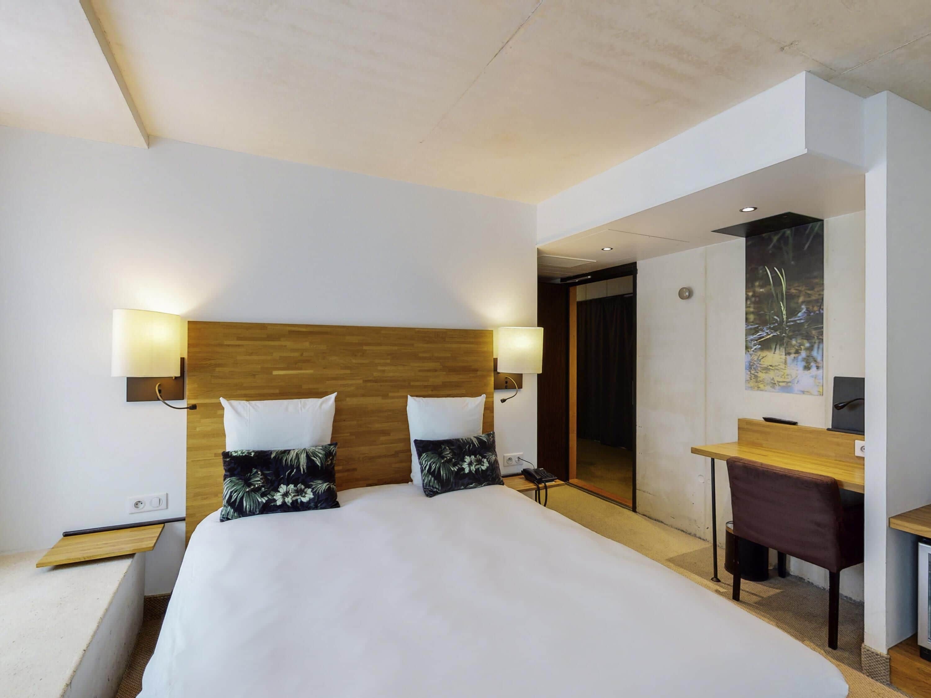 A modern hotel room with a large bed, two bedside lamps, a wooden headboard, a desk with a chair, and a painting on the wall.