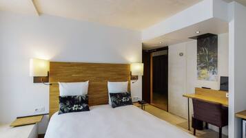 A modern hotel room with a large bed, two bedside lamps, a wooden headboard, a desk with a chair, and a painting on the wall.