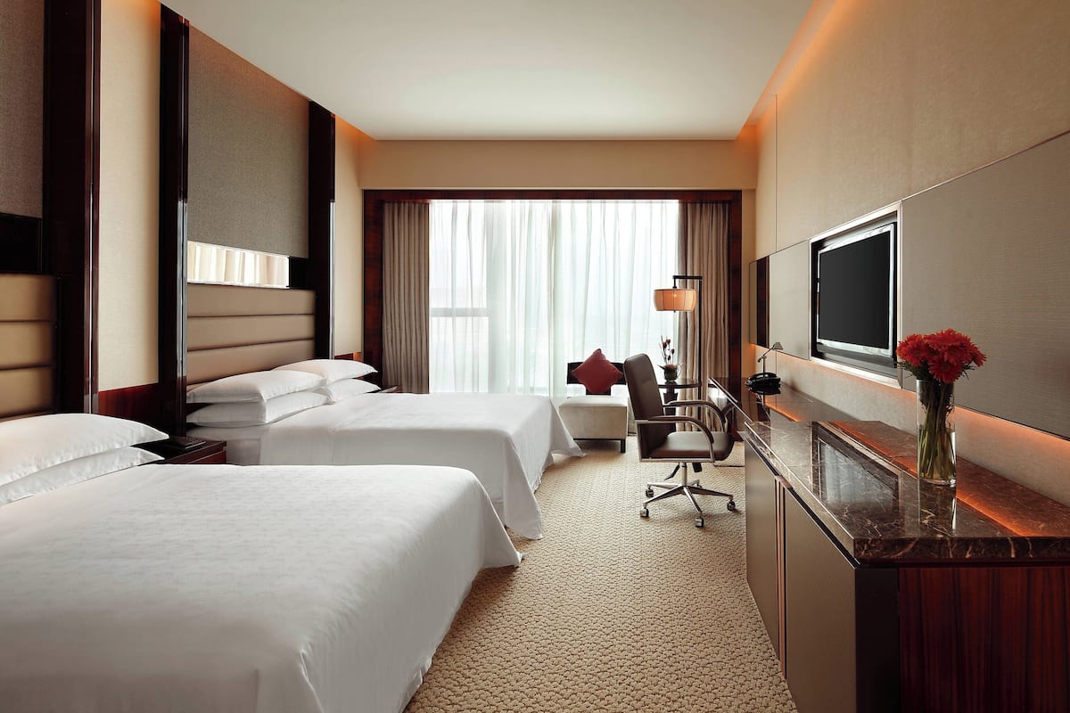A hotel room with two beds, a desk, a chair, a TV, and a vase with flowers.