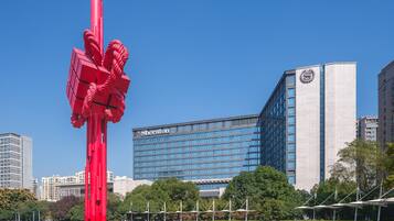Un hotel moderno con una llamativa escultura roja en primer plano, rodeado de vegetación cuidada y un cielo azul despejado.