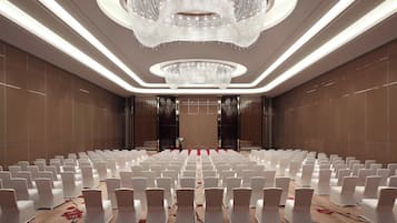 Un salón amplio con filas de sillas blancas, una lámpara de araña grande y una alfombra estampada con motivos florales.