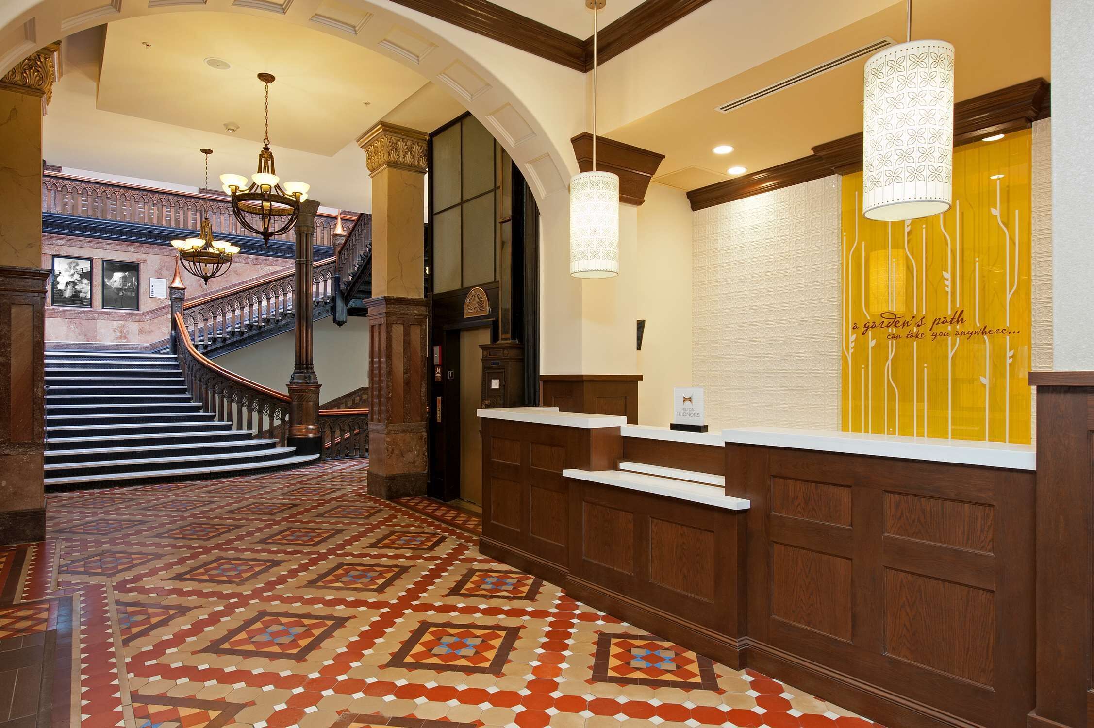 A hotel lobby with a patterned floor, a reception desk, a staircase, and a chandelier.
