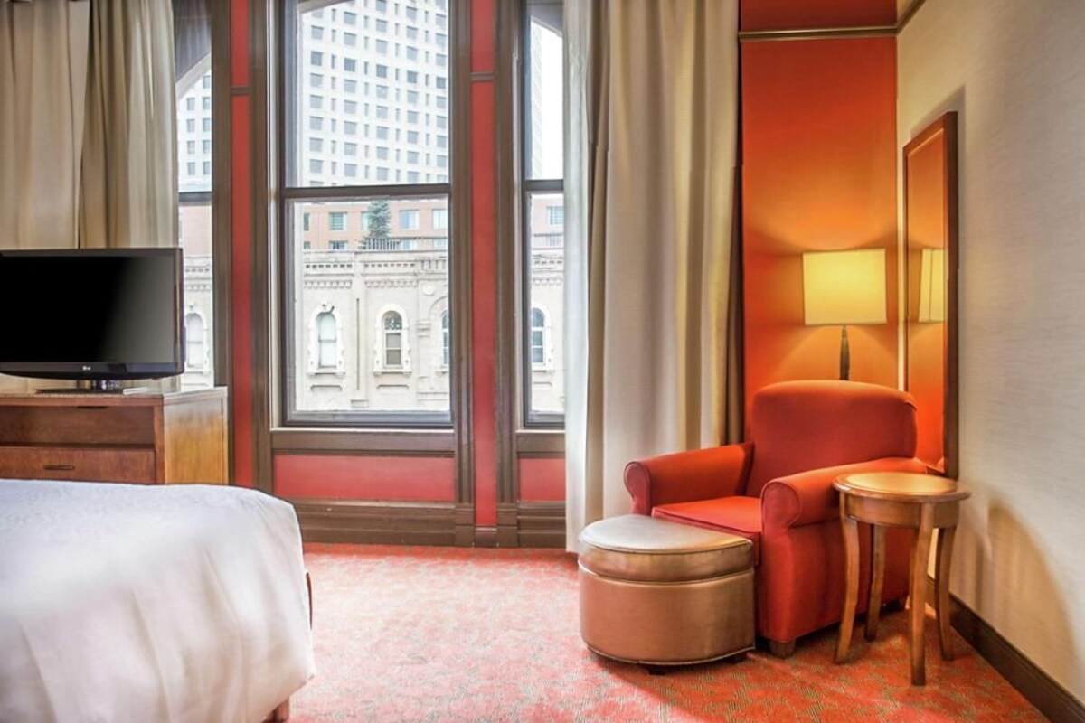 A hotel room with a bed, a television, a red armchair, a small table, and a lamp.