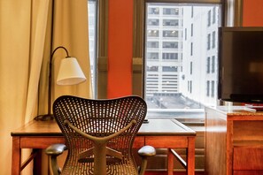 A hotel room with a wooden desk, a metal chair, a lamp, a television, and a window with a city view. - Hilton Garden Inn Milwaukee Downtown (Milwaukee)