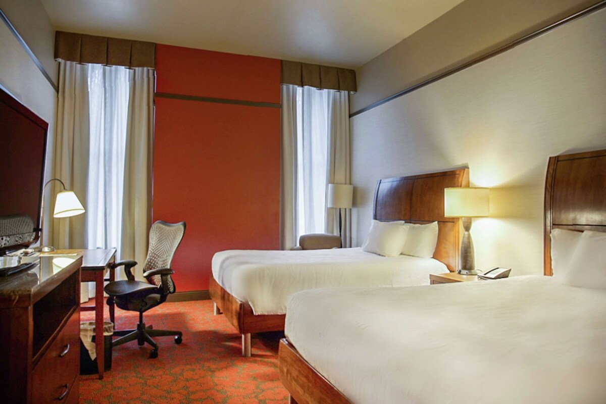 A hotel room with two beds, a desk with a chair, a lamp, and a television.