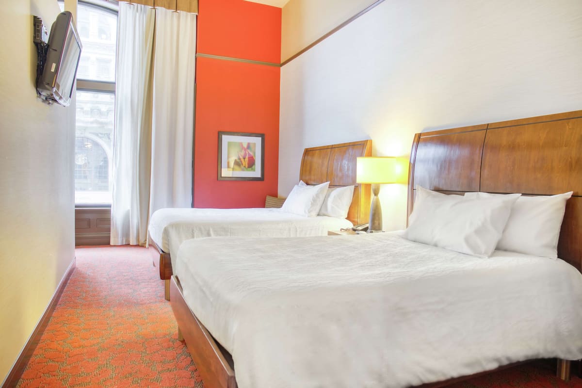 Una habitación de hotel con dos camas, una ventana con cortinas, un cuadro enmarcado en la pared y una lámpara en la mesita de noche.