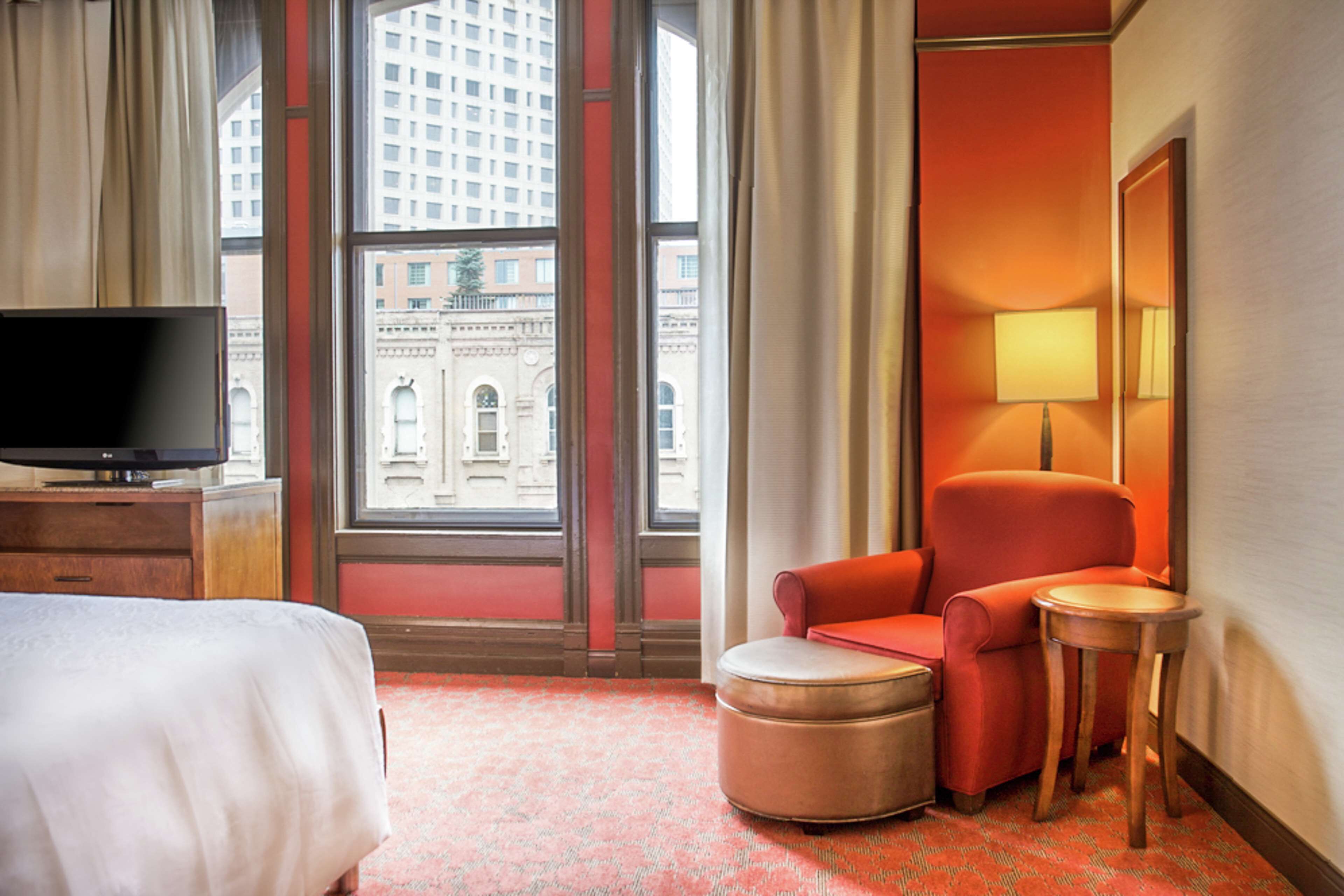 Ein Hotelzimmer mit einem Bett, einem Fernseher, einem roten Sessel, einem kleinen Hocker, einer Lampe und einem Fenster mit Blick auf ein Gebäude.