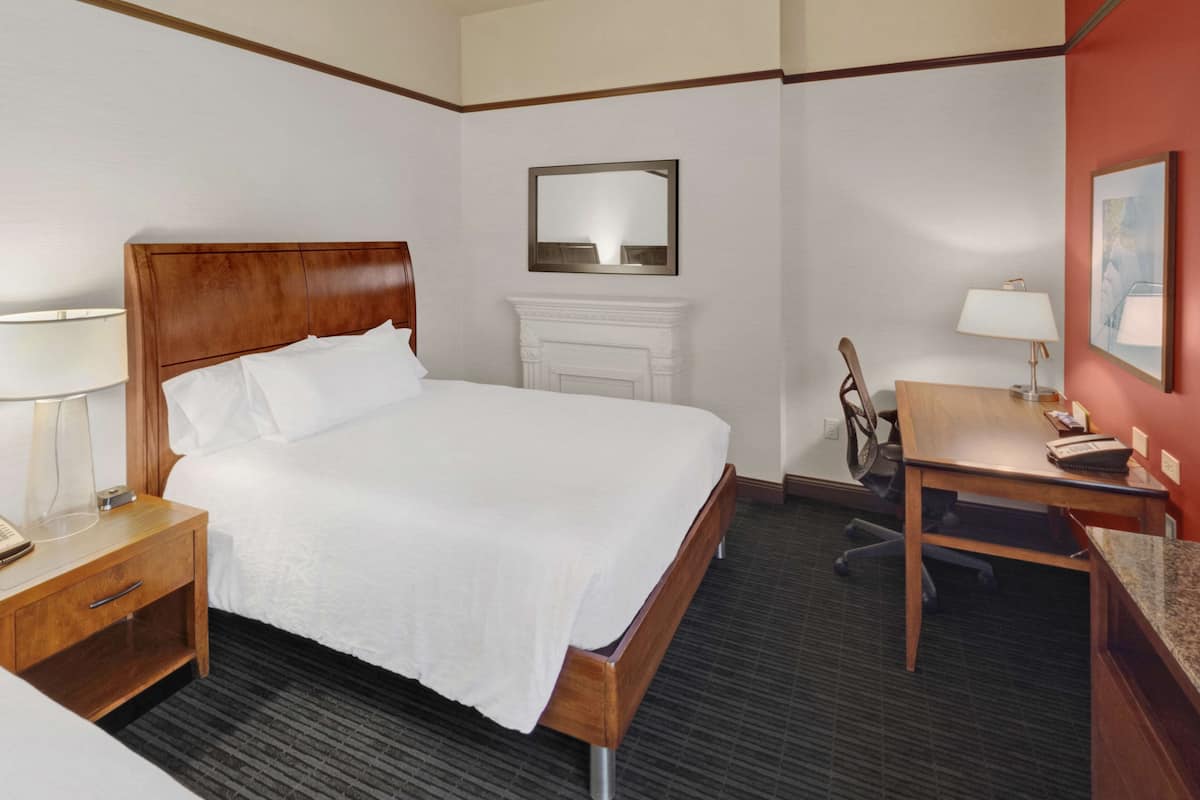 Habitación de hotel con una cama de madera, un escritorio con una silla, una mesita de noche, una lámpara y un espejo.