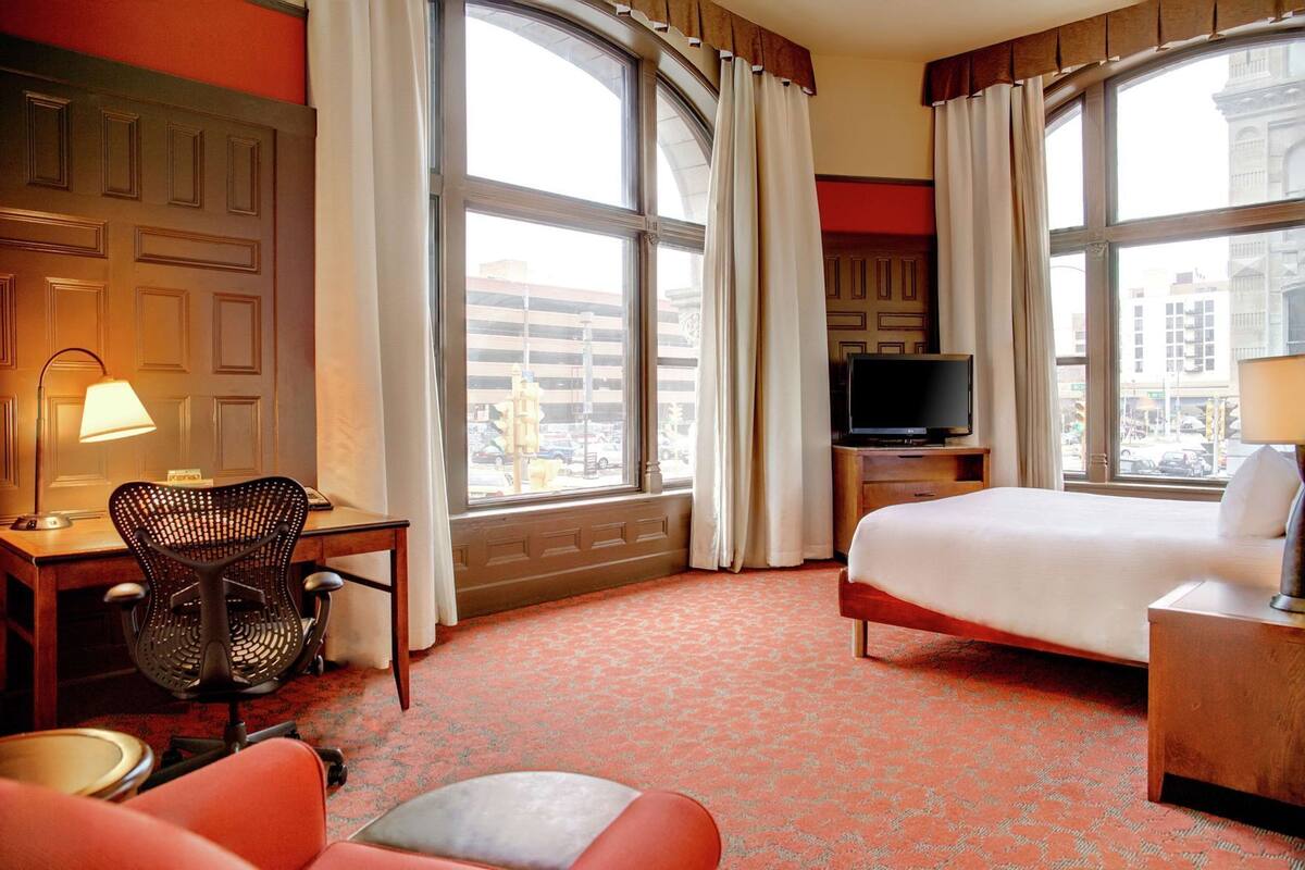 Habitación de hotel con cama, escritorio, silla, televisor y ventanas amplias.