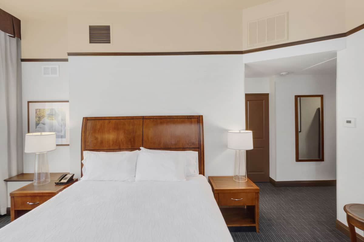 Habitación de hotel con una cama grande, dos mesitas de noche con lámparas, un cabecero de madera, un cuadro en la pared y una puerta que da a otra habitación.