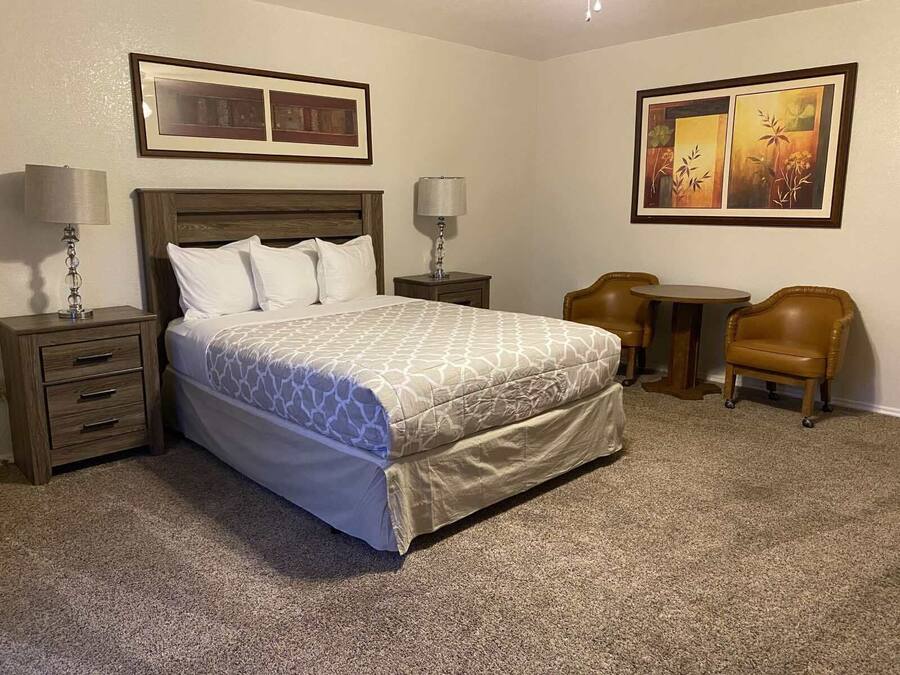Una camera d'albergo con un letto, due comodini, una lampada, una sedia, un tavolino e due quadri appesi al muro.