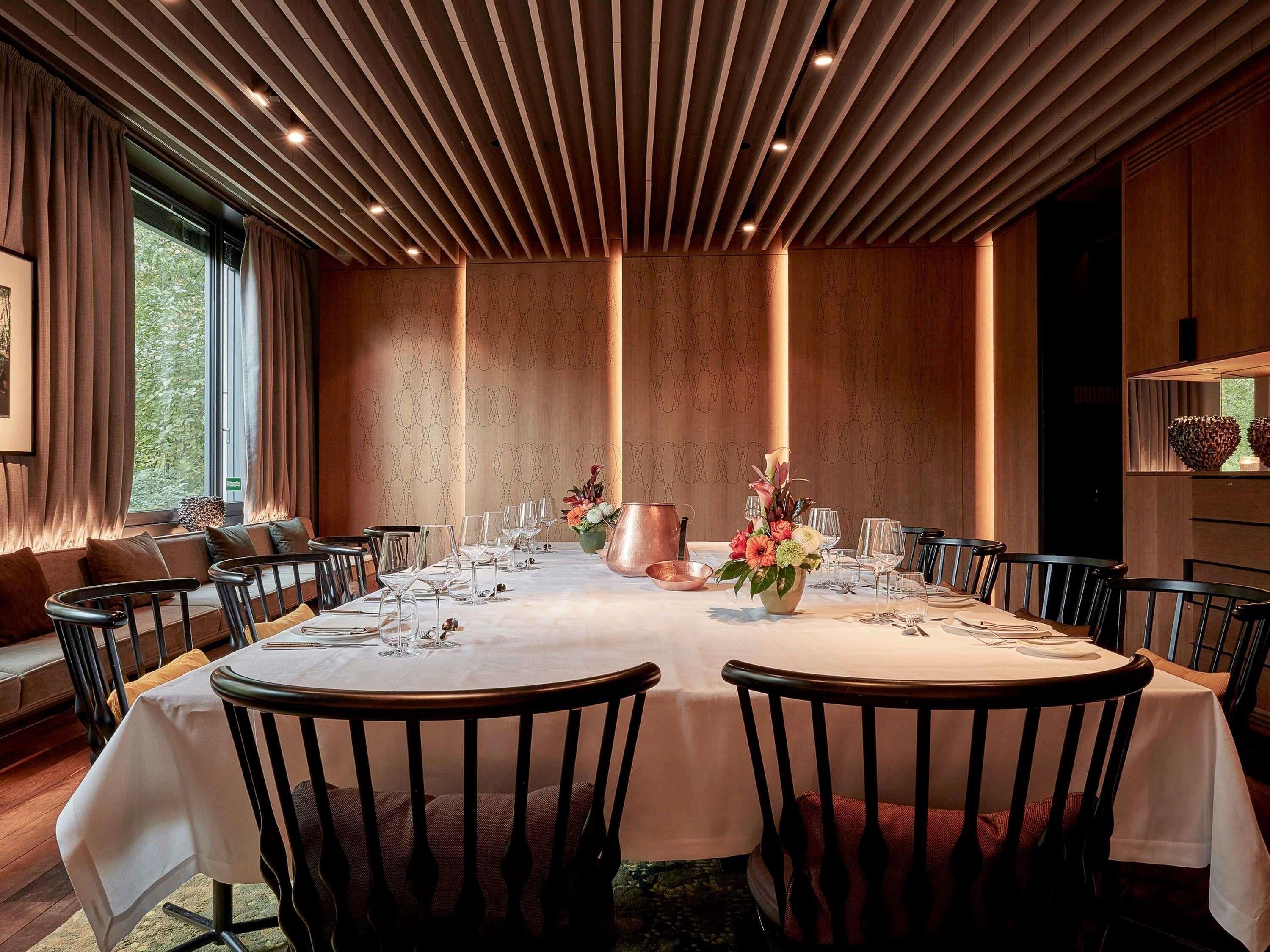 A dining room with a long table set for a meal, surrounded by chairs, with a patterned wall and a window with curtains.