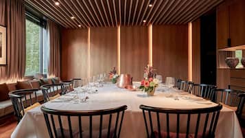 A dining room with a long table set for a meal, surrounded by chairs, with a patterned wall and a window with curtains.