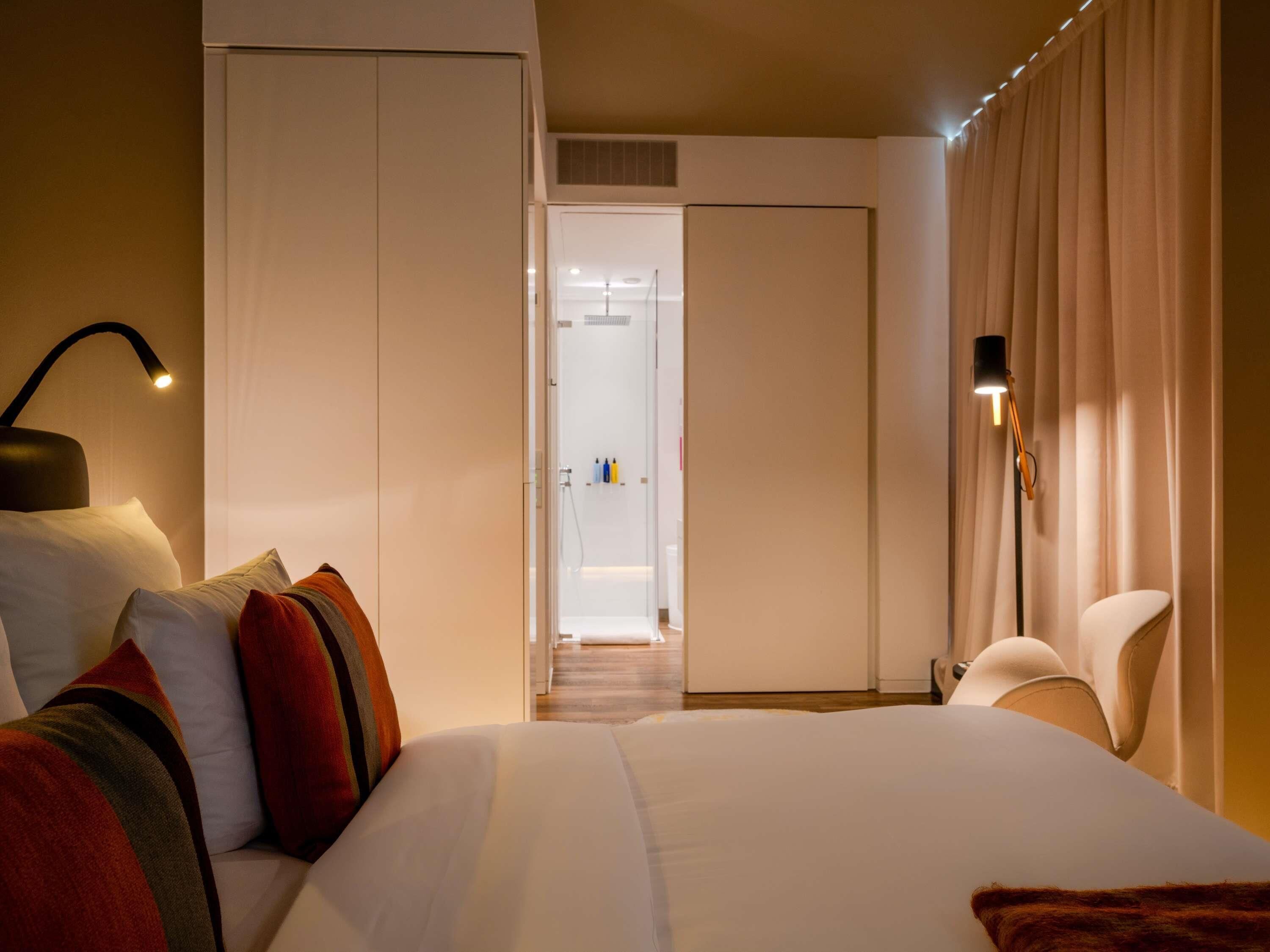 Habitación de hotel con cama, lámparas de noche, armario y vista al baño.