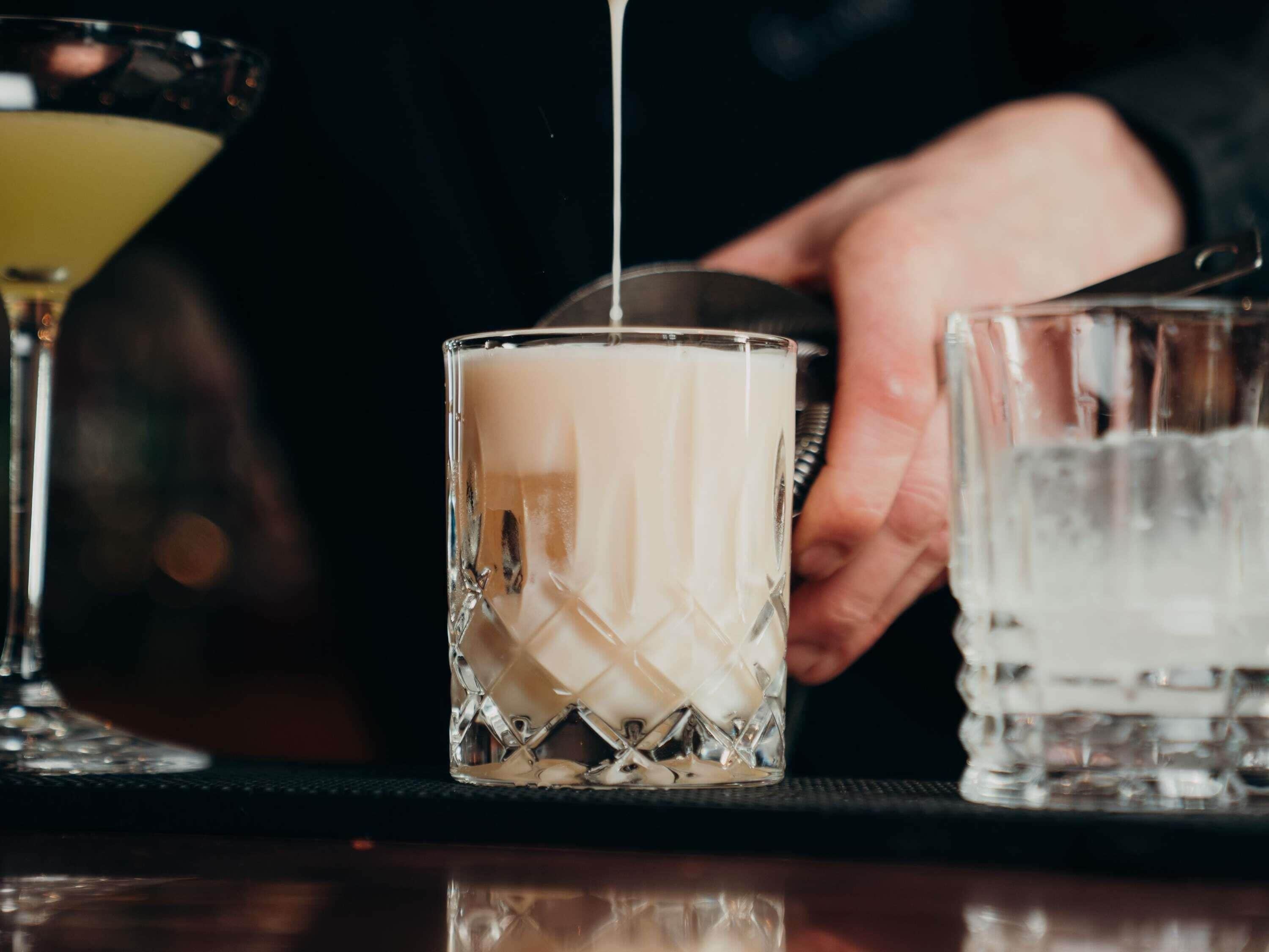A bar setting with a glass being filled with a white liquid, likely milk or cream, by a bartender.