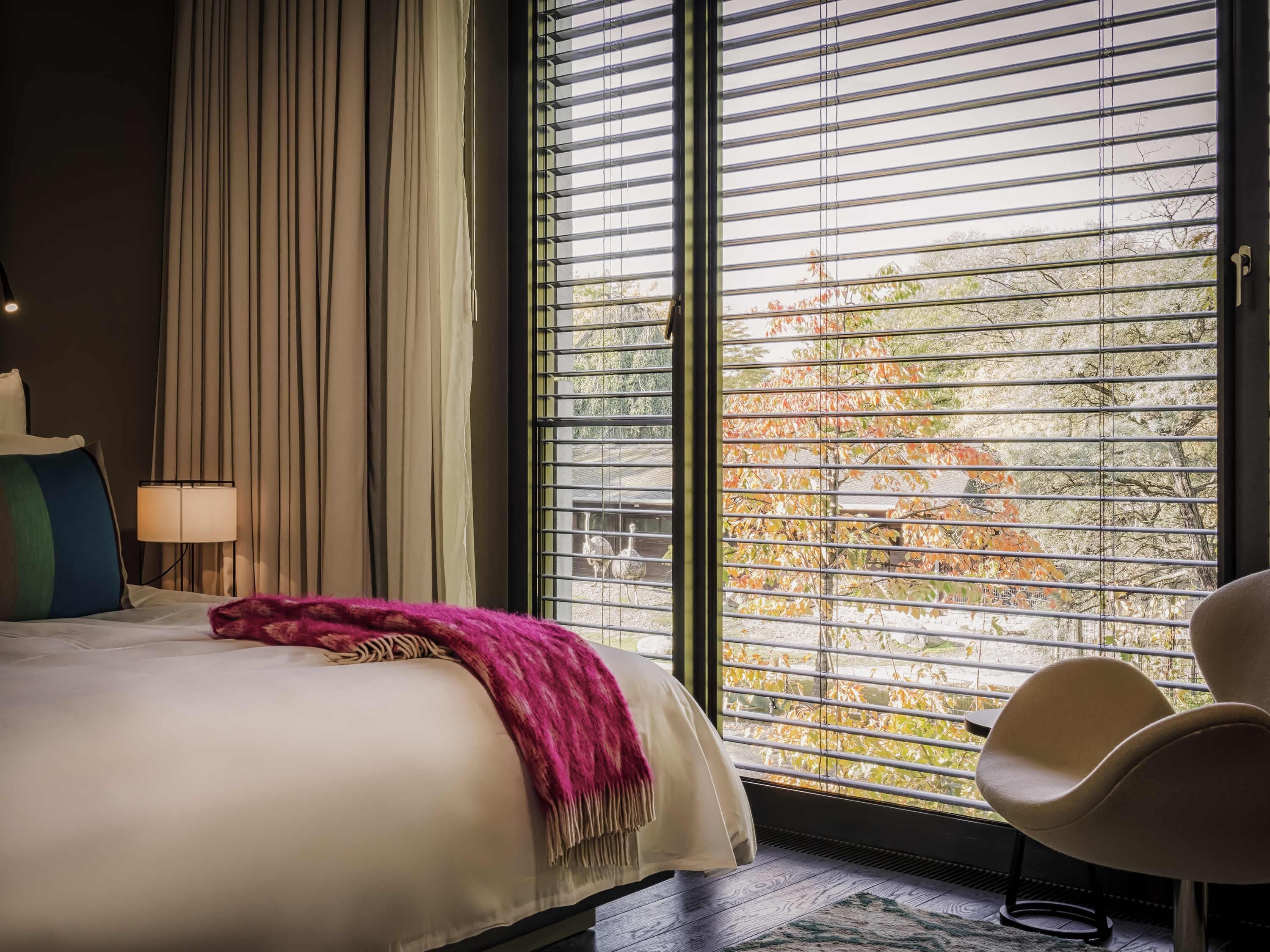 Une chambre d’hôtel avec un lit, une chaise, une fenêtre avec des stores et une lampe.