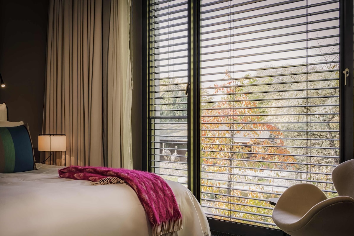 A hotel room with a bed, a chair, a window with blinds, and a lamp.