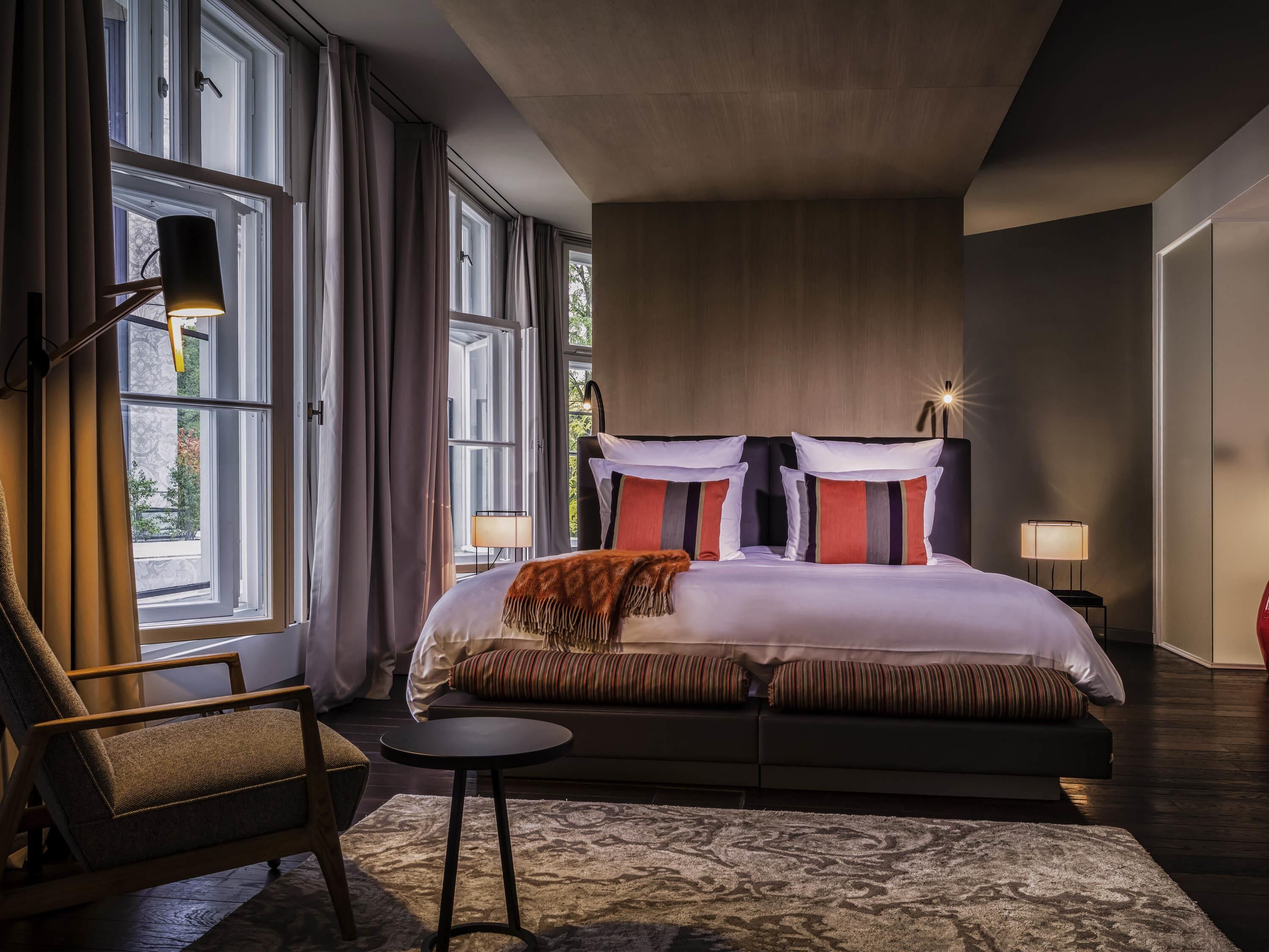 Una habitación de hotel moderna con una cama grande, una silla, una mesita y varios ventanales.