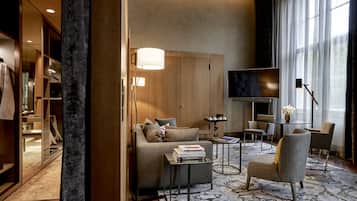 Una habitación de hotel moderna con sofá, sillones, una mesa de centro, televisor y un amplio ventanal con cortinas.