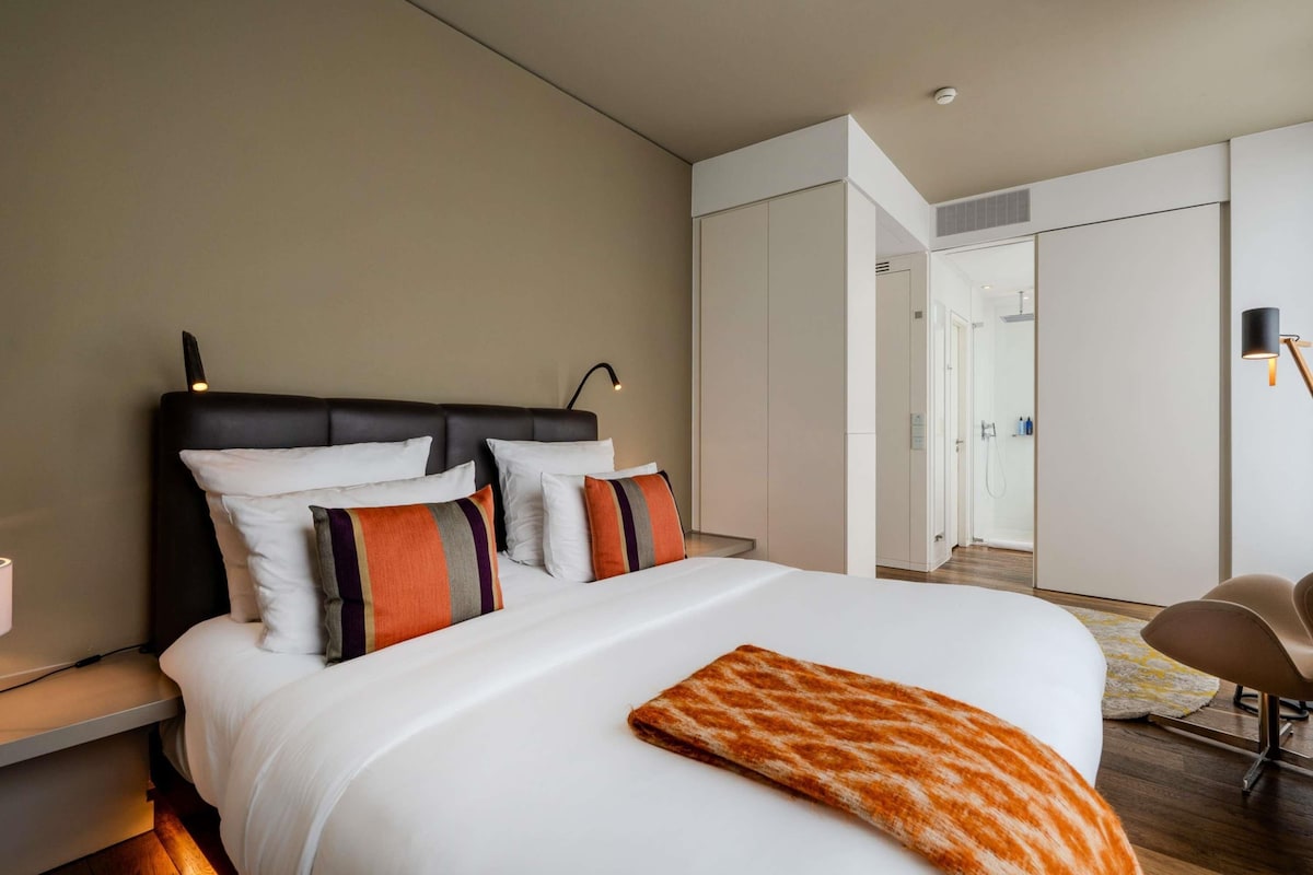 A modern hotel room with a large bed, a bedside table, a chair, and a walk-in closet.
