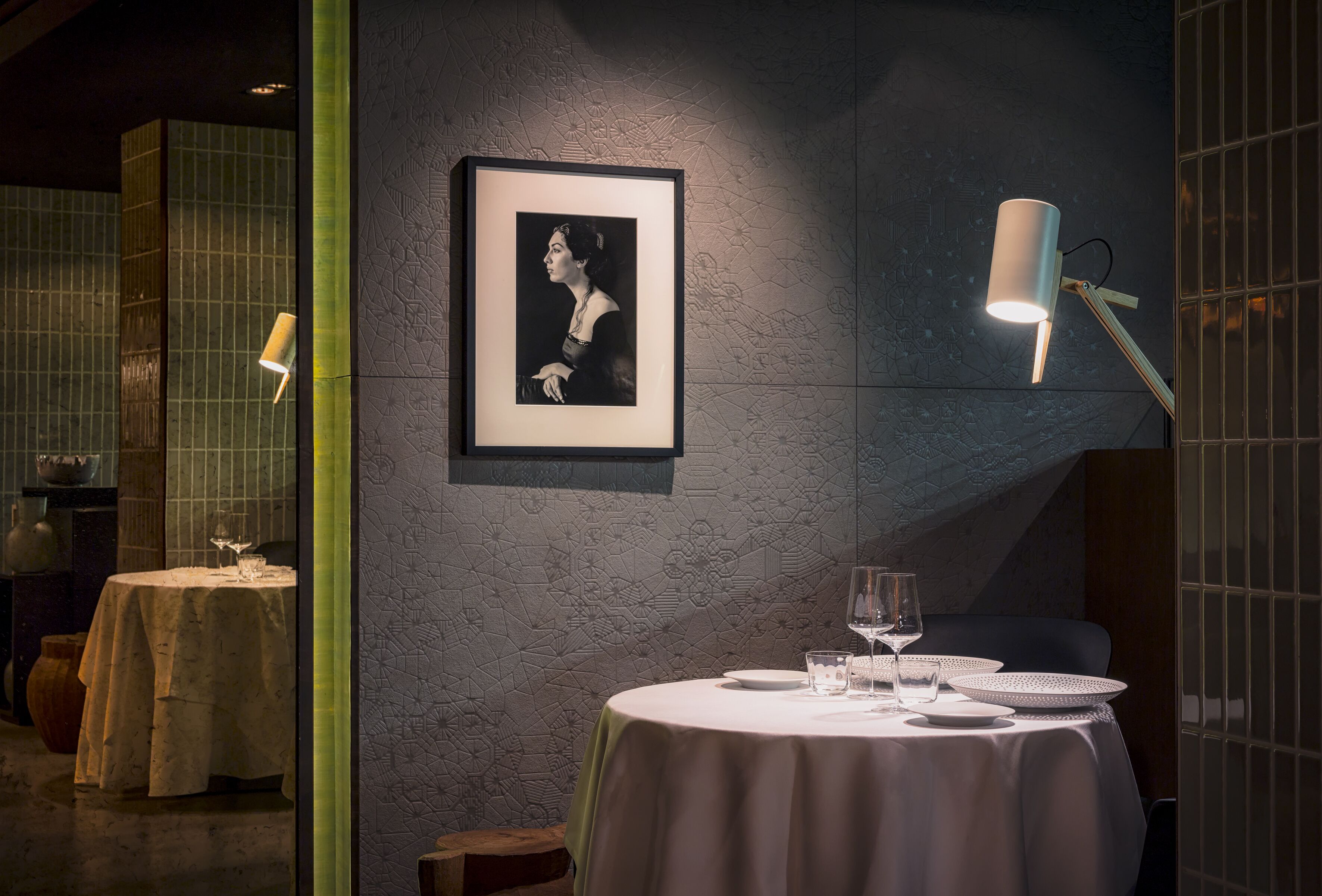 A dining area with a round table set for two, a framed portrait on the wall, and a wall-mounted lamp.
