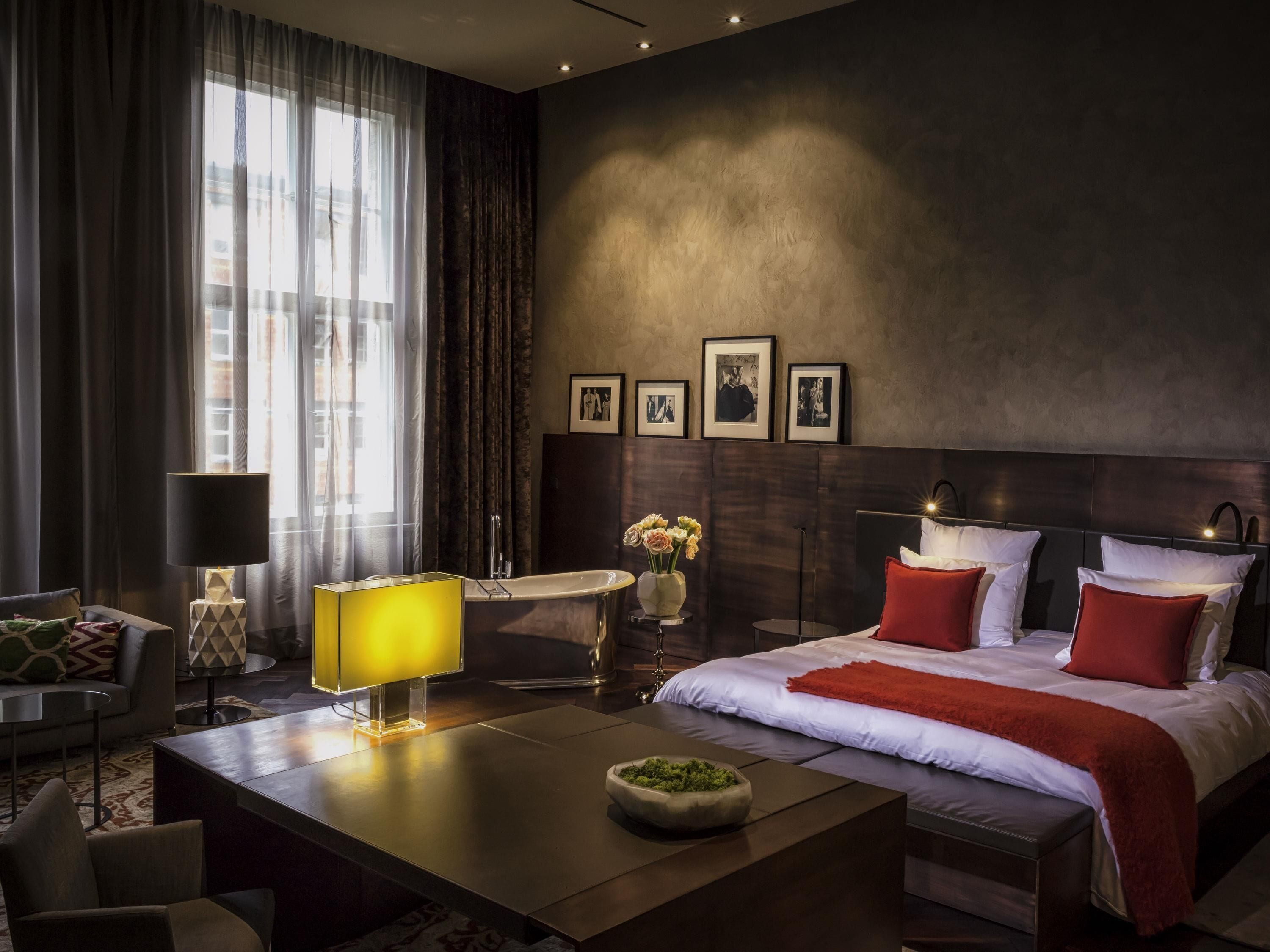 A modern hotel room with a large bed, a desk with a lamp, a vanity with a mirror, and a window with curtains.