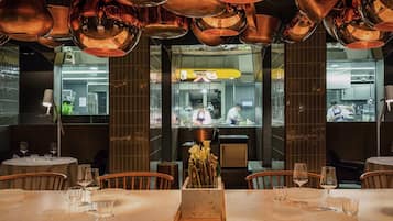 A restaurant interior with a long table set for dining, a kitchen area visible through a glass partition, and copper fixtures on the ceiling.