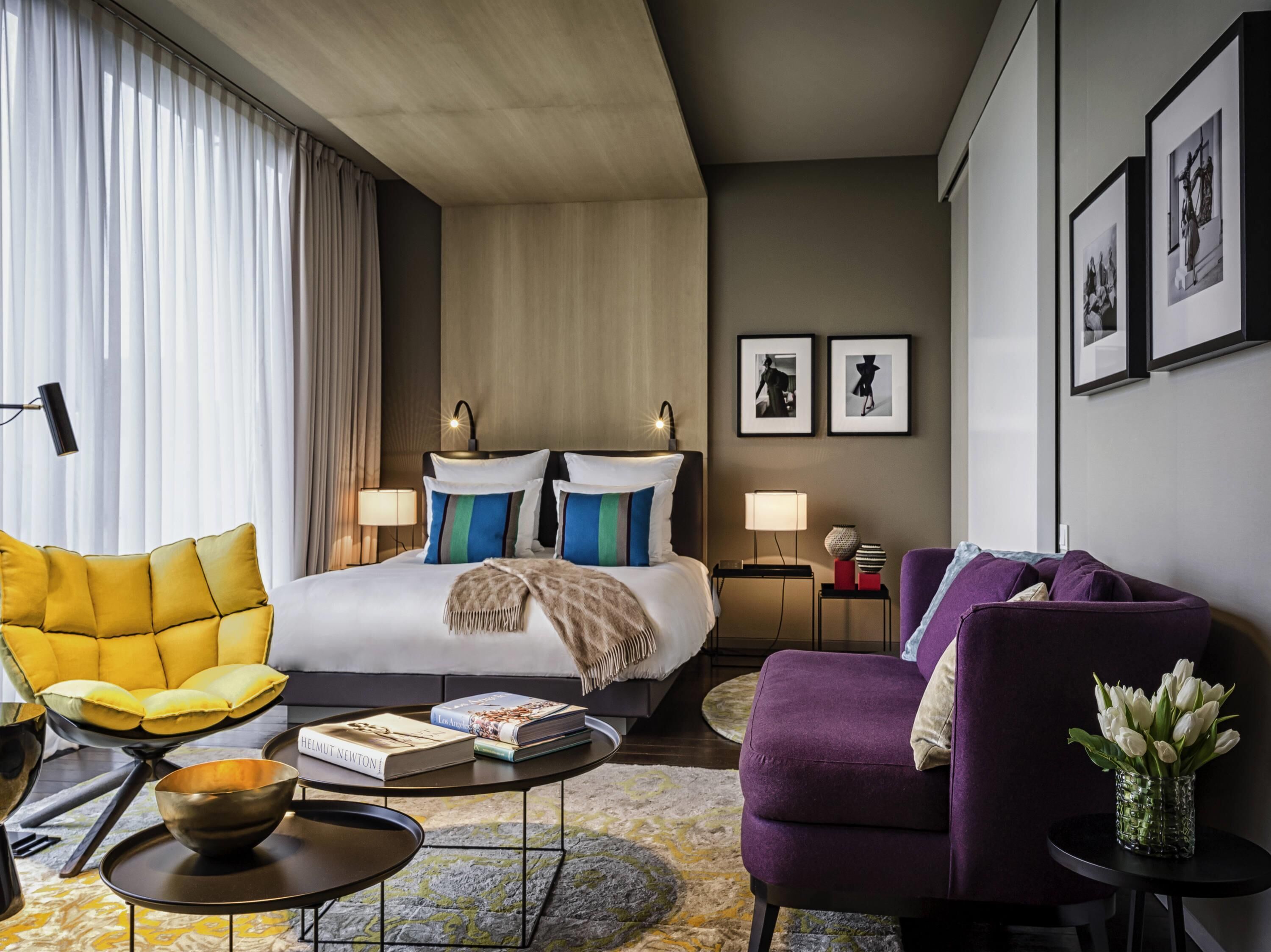 Habitación de hotel con una cama, dos mesitas de noche, un sofá, una silla, una mesa de centro y un jarrón con flores.