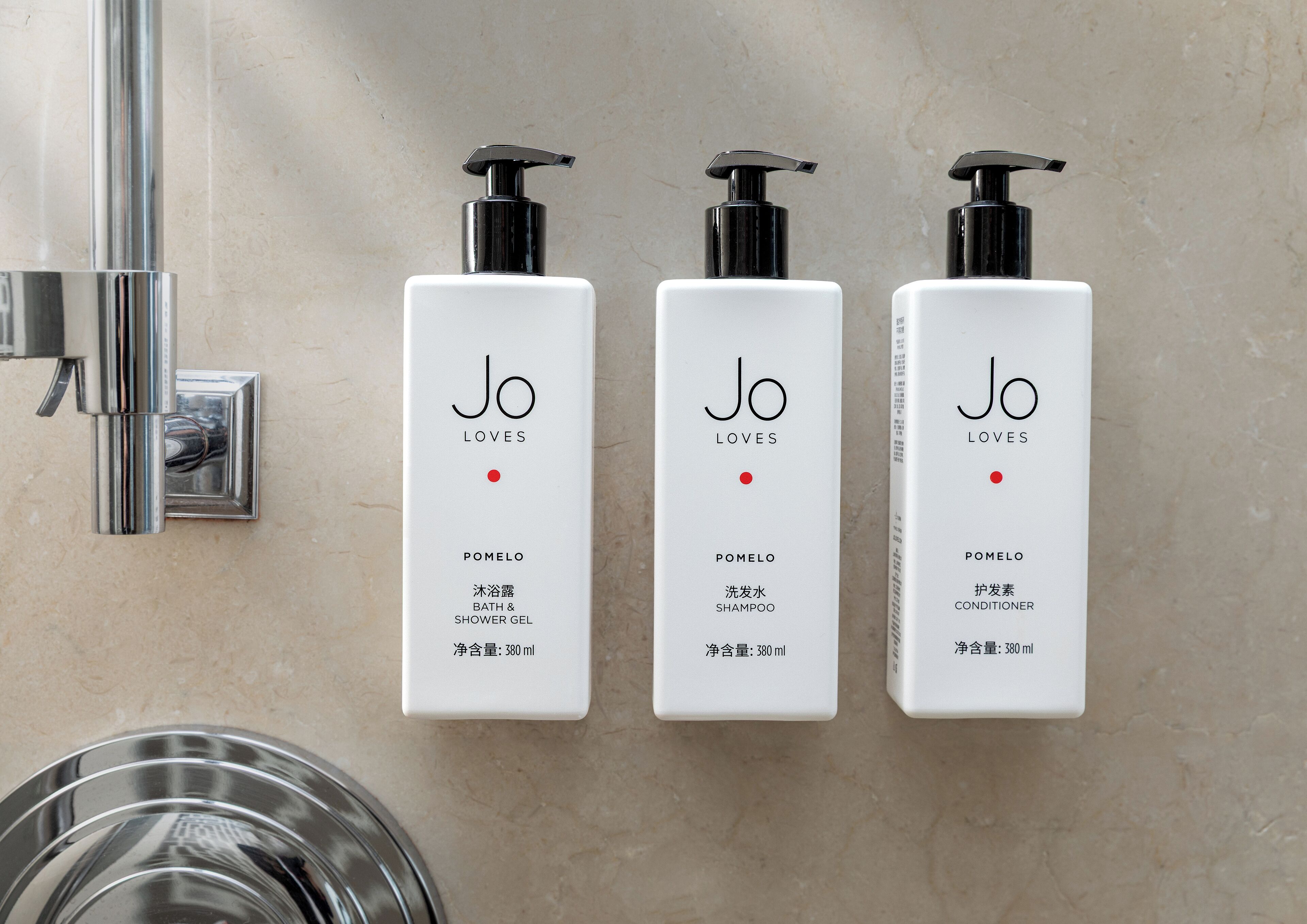 Tres botellas blancas con dosificadores negros etiquetadas 'Jo Loves' sobre una pared color beige.