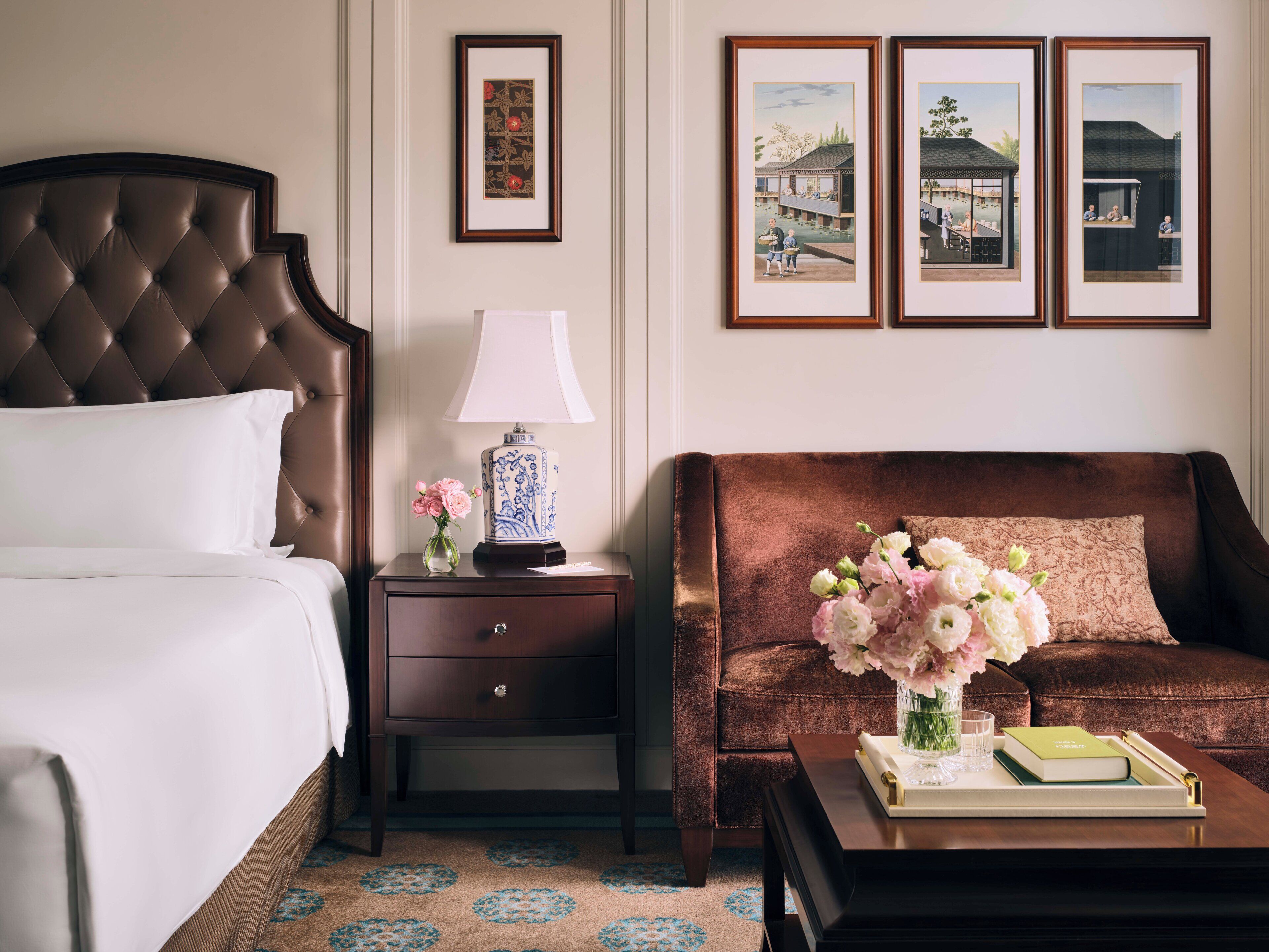 A hotel room with a tufted headboard bed, a nightstand with a lamp and vase, a sofa with a cushion, and a coffee table with a book and vase.