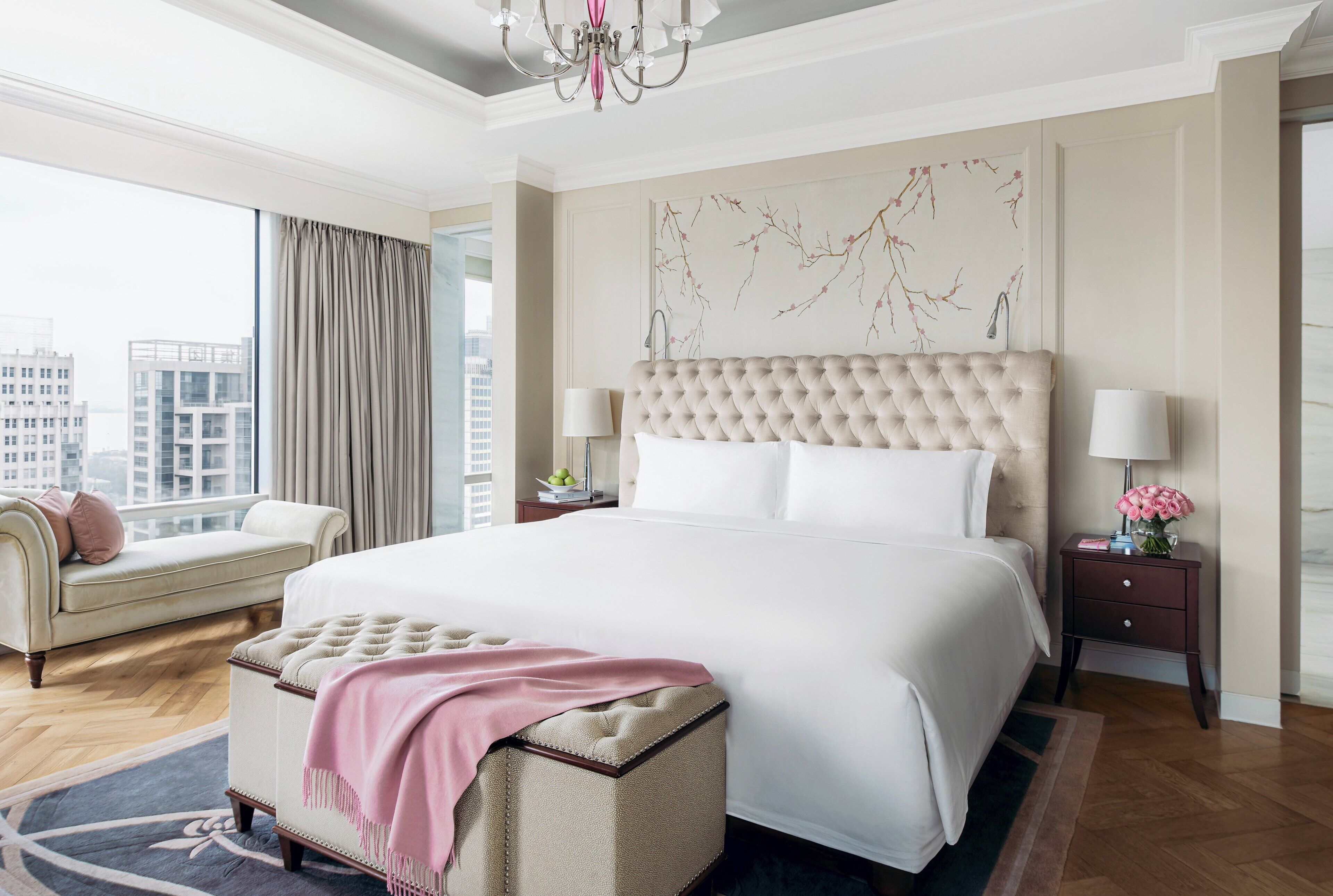 A spacious bedroom with a large bed, a chaise lounge, a nightstand with a lamp, and a view of the city.