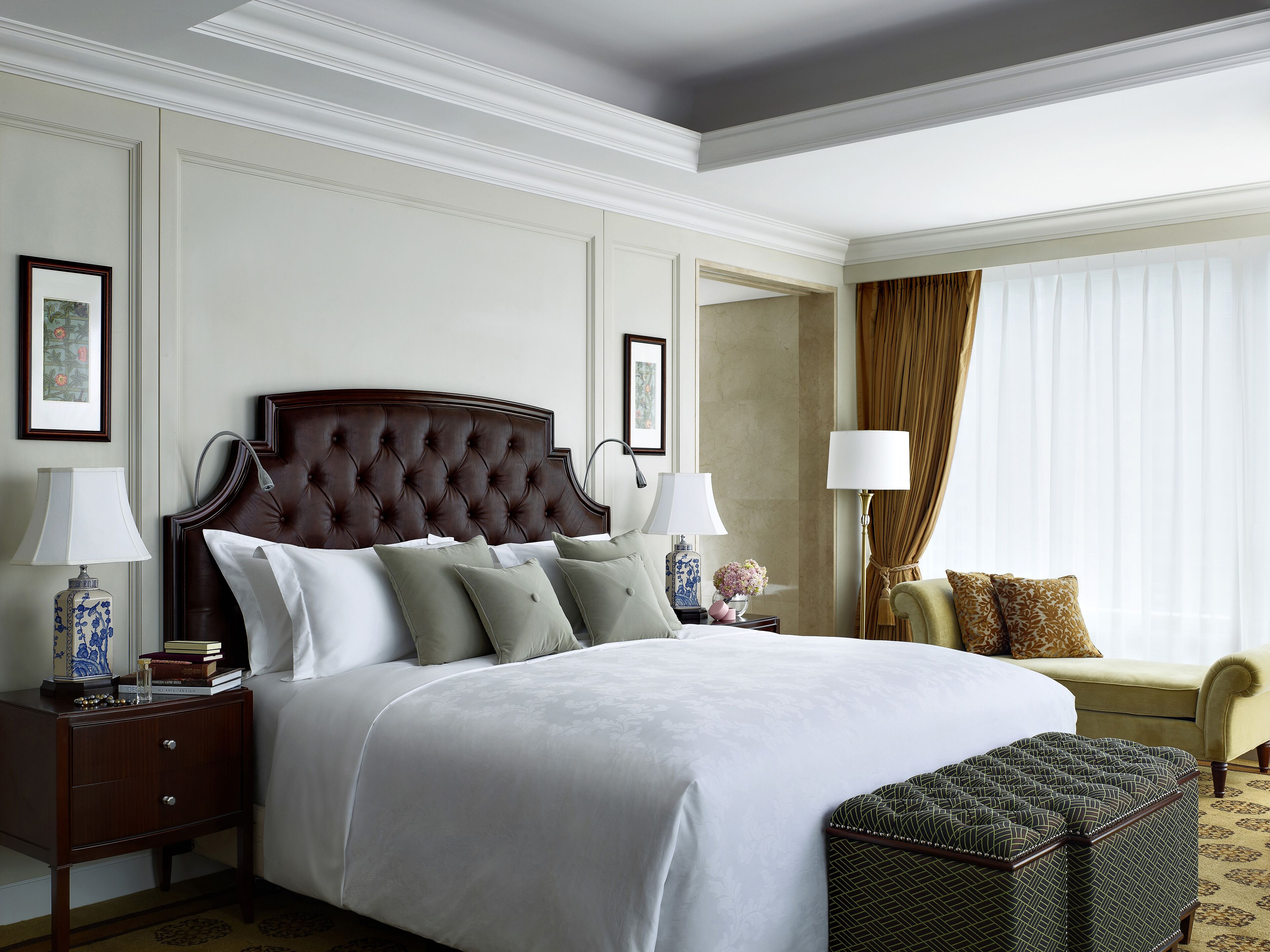 A neatly arranged hotel room with a large bed, a bedside table with a lamp, a small table with a vase, a chair, and a patterned ottoman.
