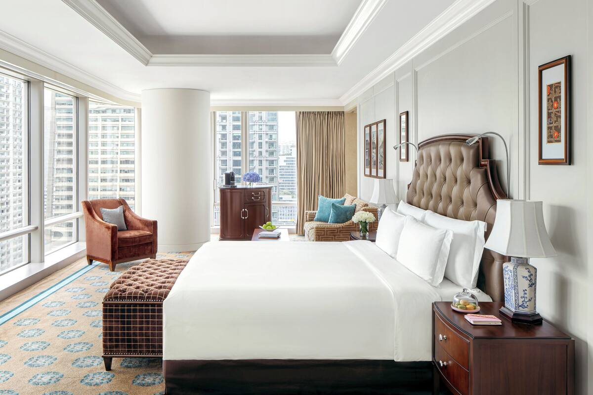 Ein Hotelzimmer mit einem großen Bett, einer Sitzecke mit einem Sessel, einem Nachttisch und Blick auf die Stadt durch große Fenster.