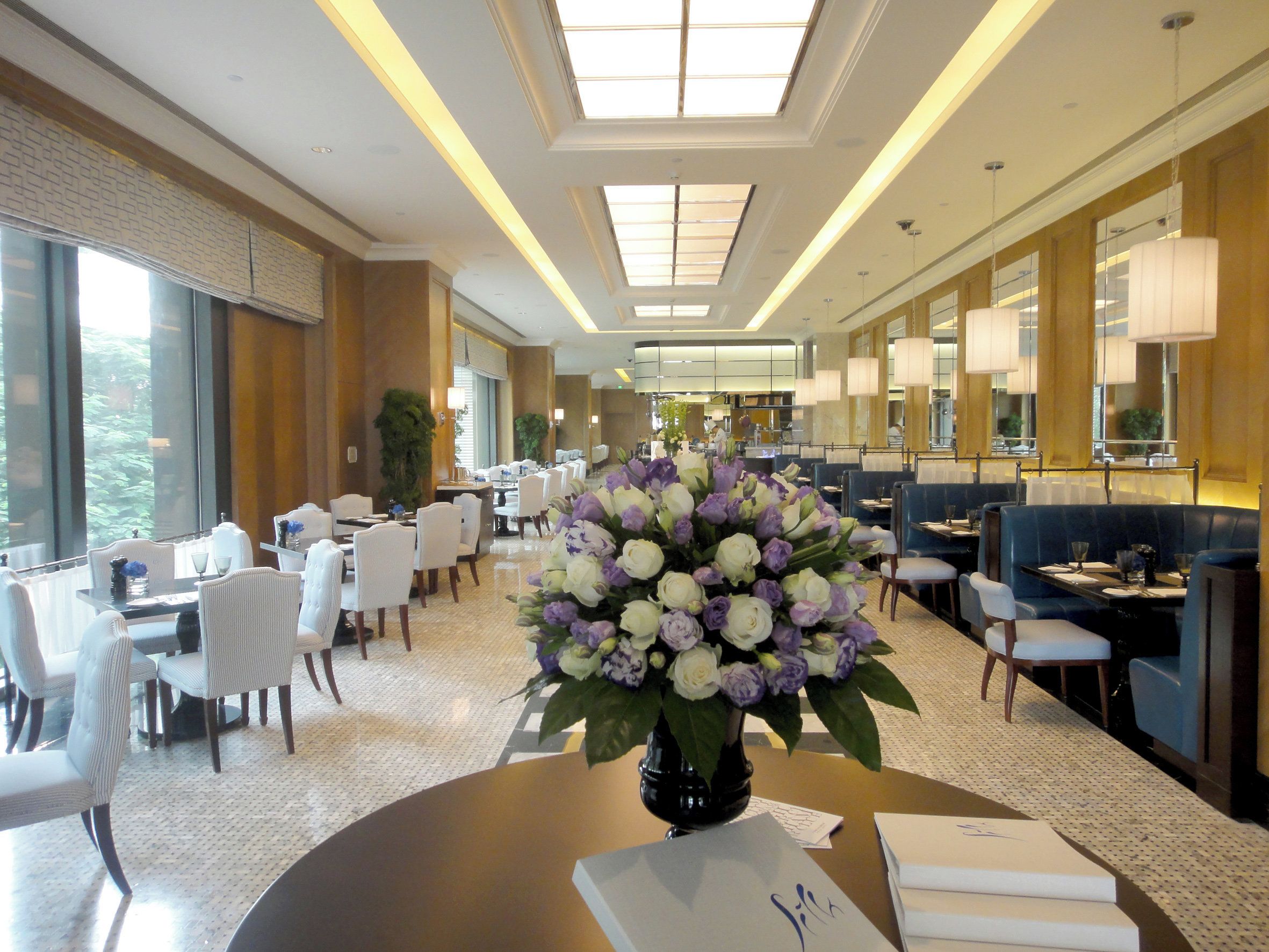 A hotel lobby with a round table in the foreground, a large floral arrangement, and a seating area with tables and chairs.