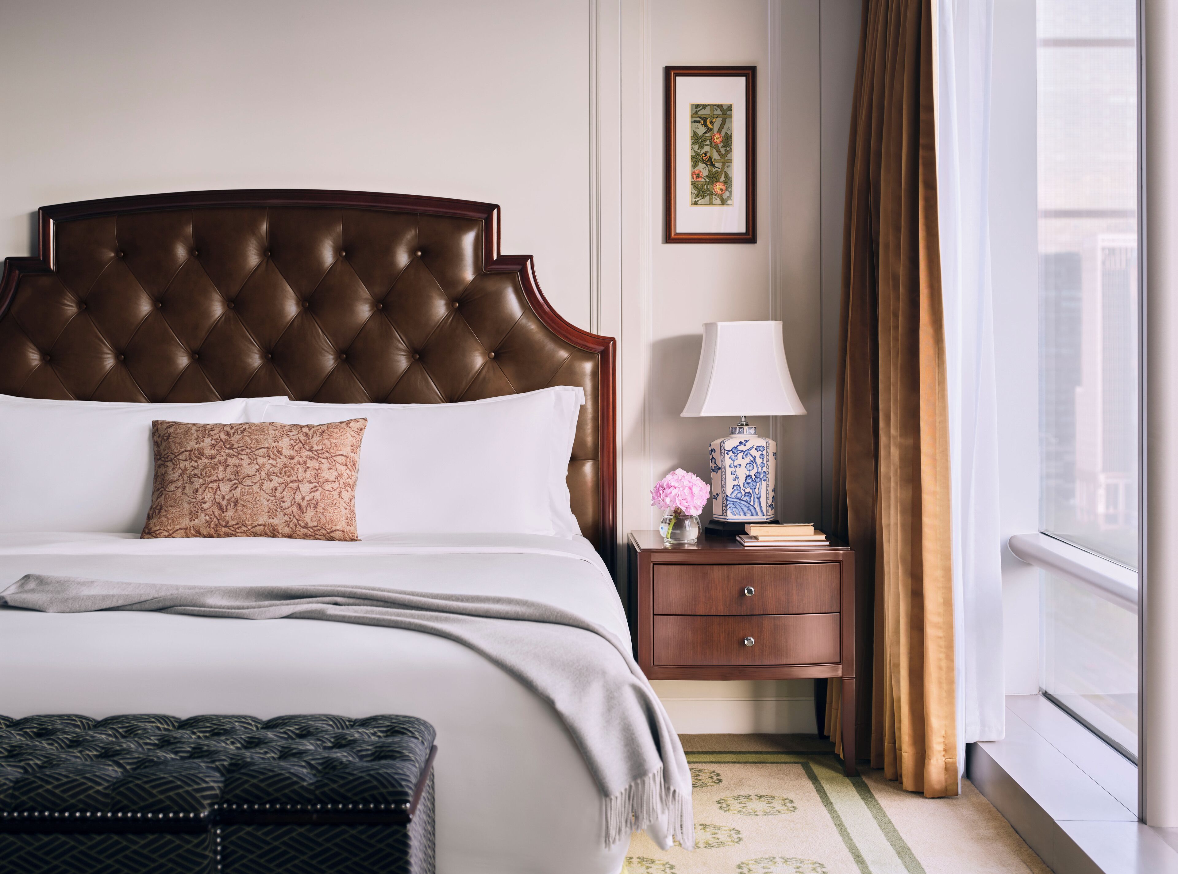 Una cama bien hecha con un cabecero capitonado, una mesita de noche con una lámpara y un jarrón de flores, y una ventana con cortinas.