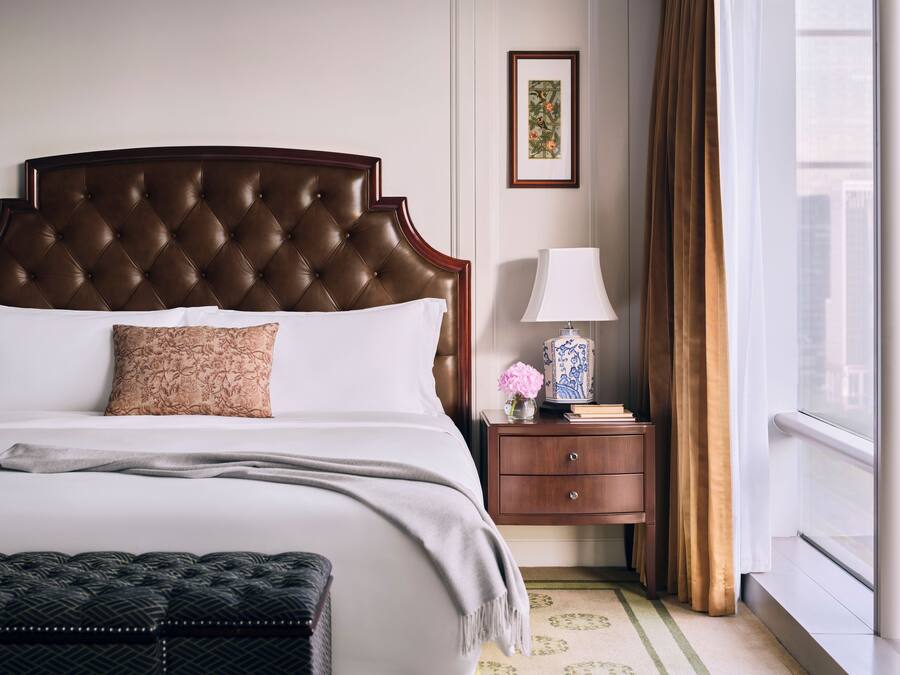 A neatly made bed with a tufted headboard, a bedside table with a lamp and a vase of flowers, and a window with curtains.