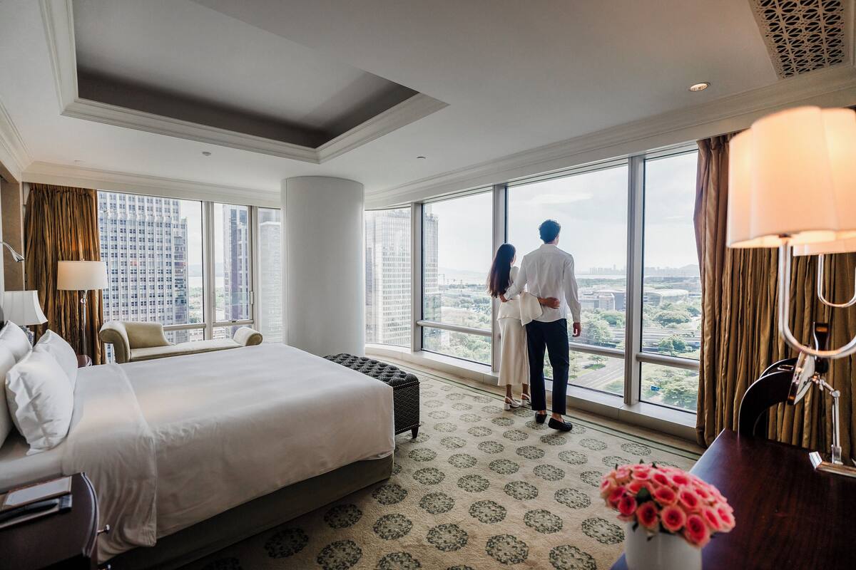 Ein Hotelzimmer mit einem großen Bett, einem Schreibtisch und Blick auf die Stadt durch große Fenster.