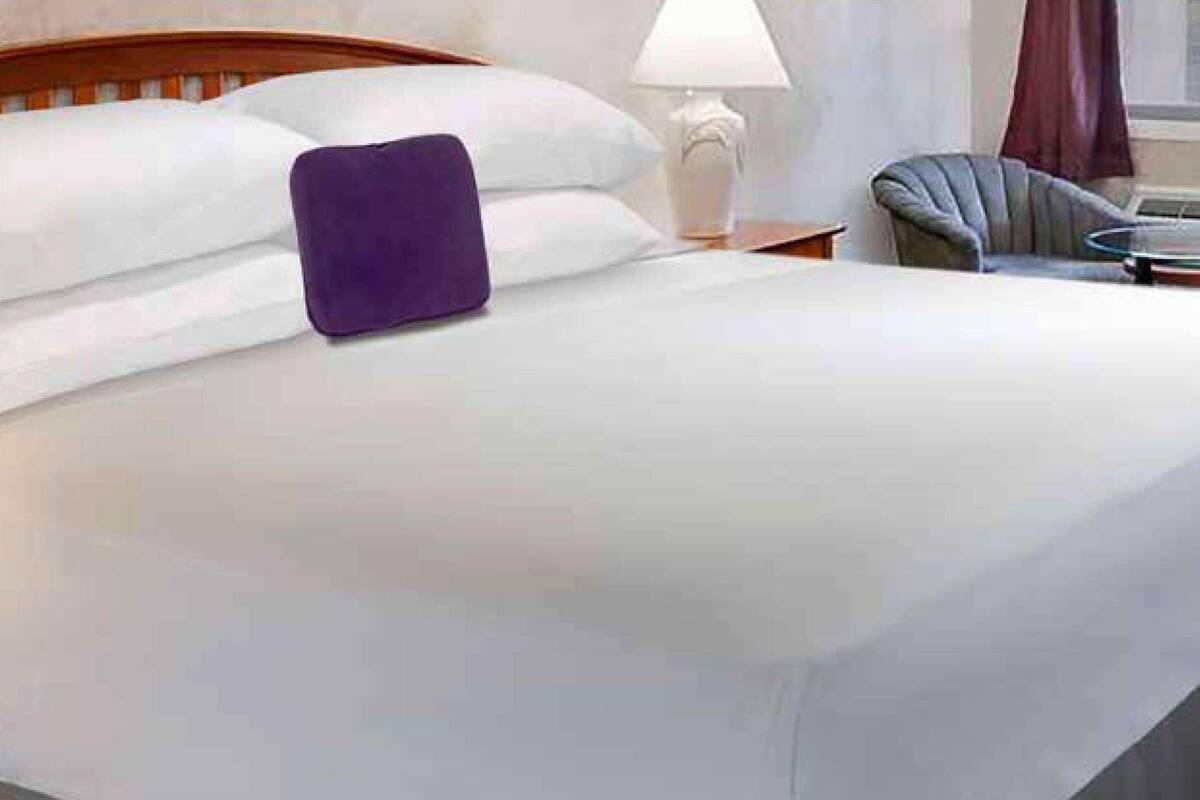 A neatly made bed with white linens and a purple pillow, a wooden headboard, a bedside table with a lamp, and a chair with a round table beside the bed.