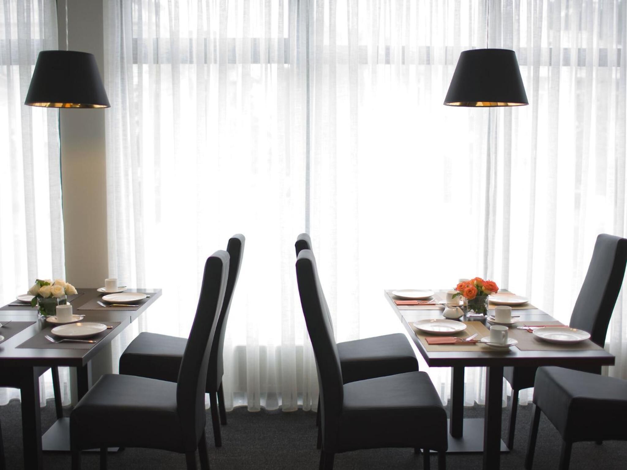 A dining area with tables set for four, each with a black chair, white plates, and a small vase of flowers.