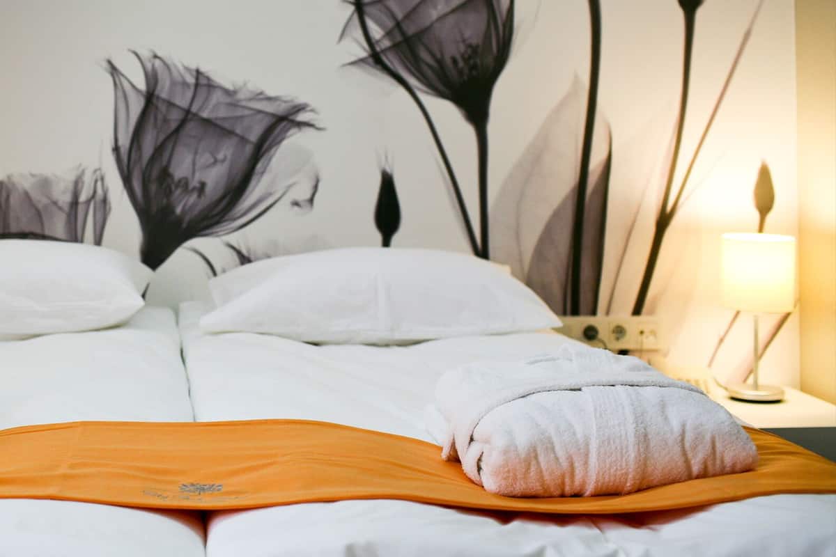 A hotel room with a bed, white bedding, a pillow, an orange blanket, and a wall with a floral mural.